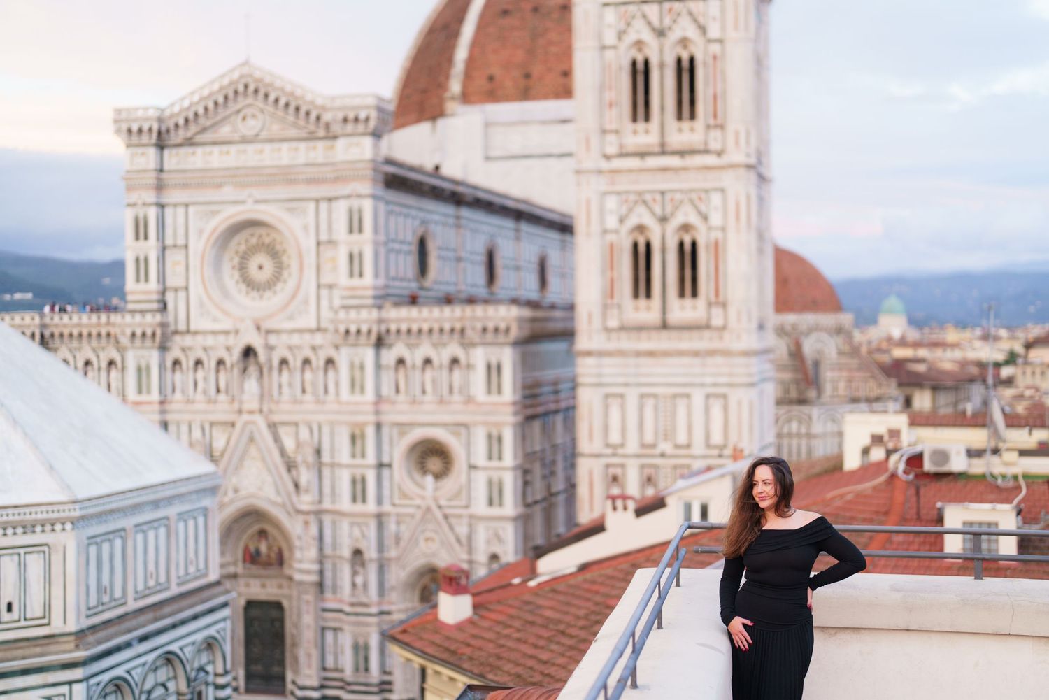 Tourist poses on rooftop terrace overlooking Florence Cathedral and Giotto's Bell Tower in Italy at sunset.