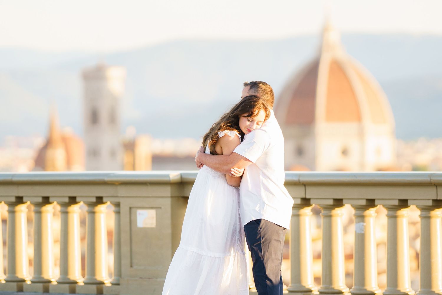 A couple in white outfits shares a romantic moment on a balcony overlooking the ocean during sunset.