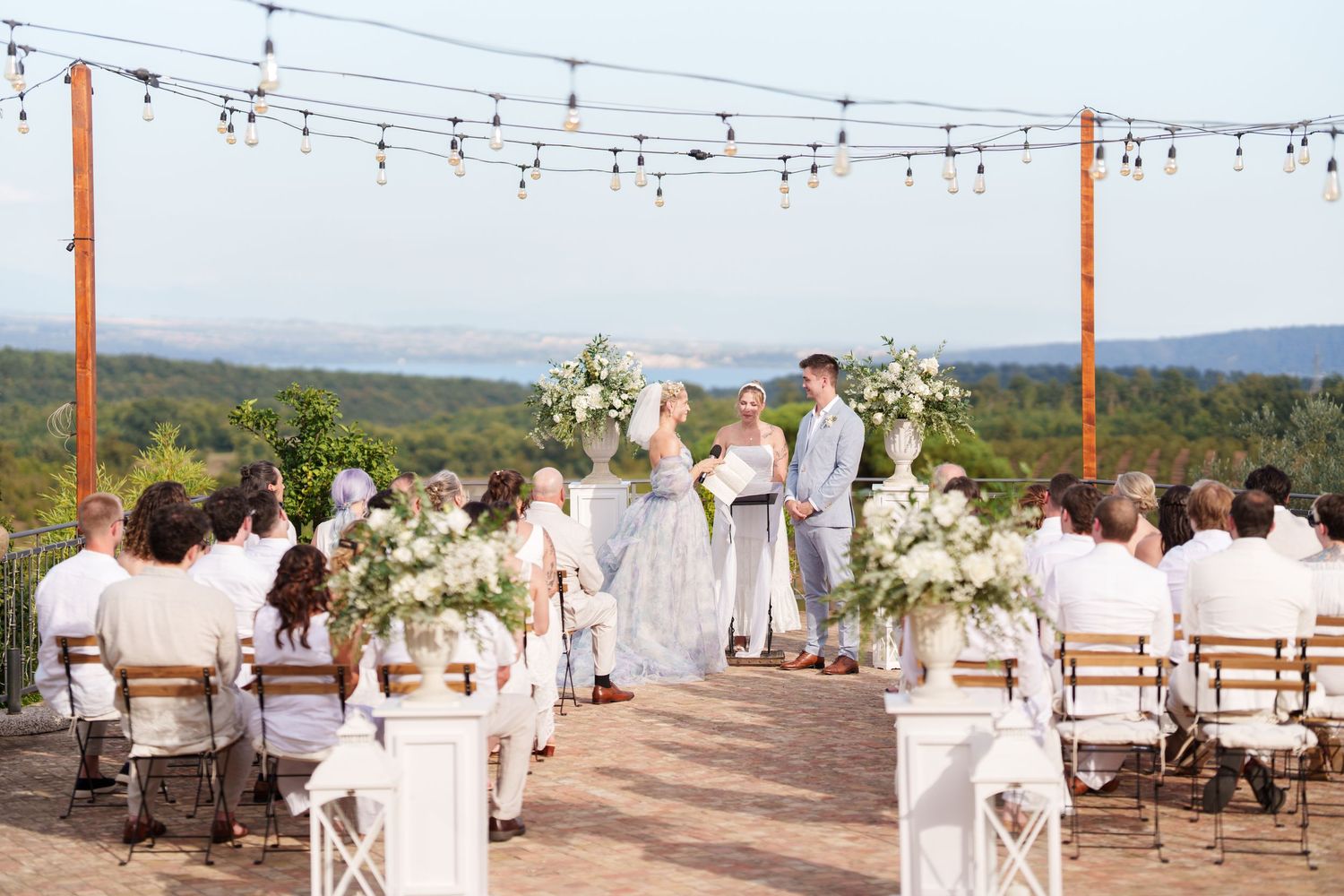 Outdoor wedding ceremony with string lights, white floral arrangements, and scenic mountain views.
