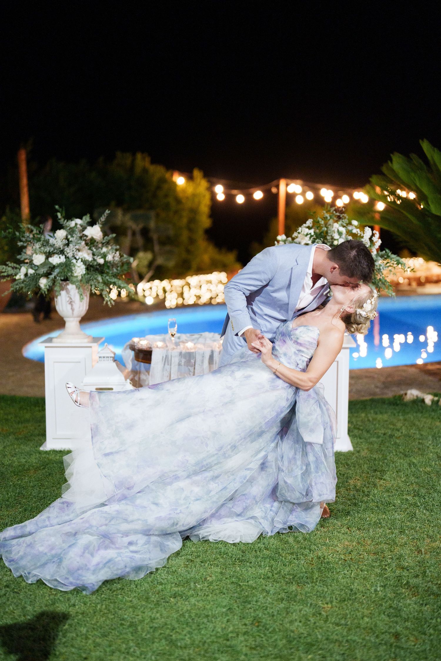 Bride in blue floral gown shares romantic dip kiss with groom in gray suit by pool at night wedding reception.