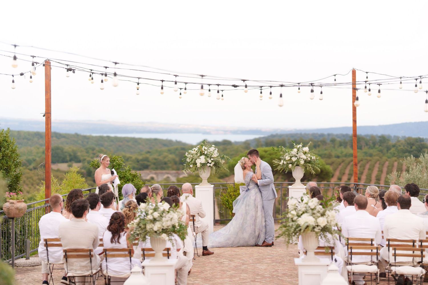 Outdoor vineyard wedding ceremony with couple at floral altar, string lights, and scenic lake views.