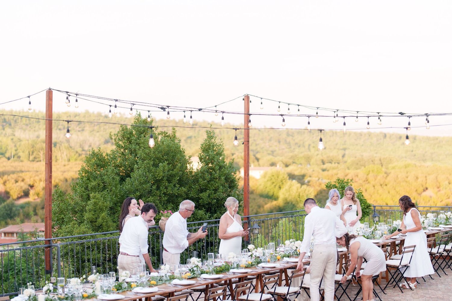 Elegant outdoor wedding reception with string lights, long white tables, and guests in white attire at golden hour.