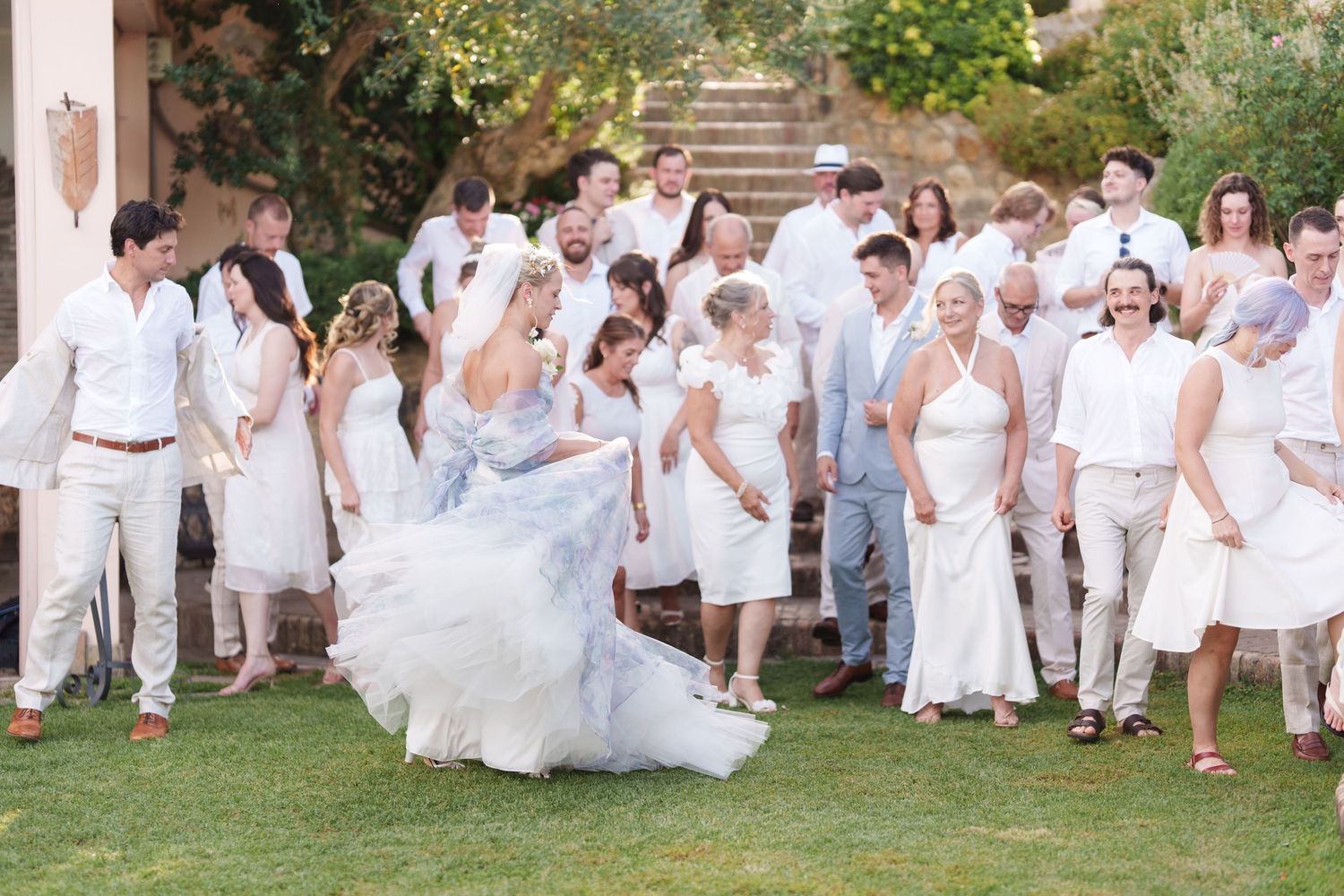 Wedding guests in white attire celebrate outdoors as bride twirls in blue-gray gown at elegant garden reception.