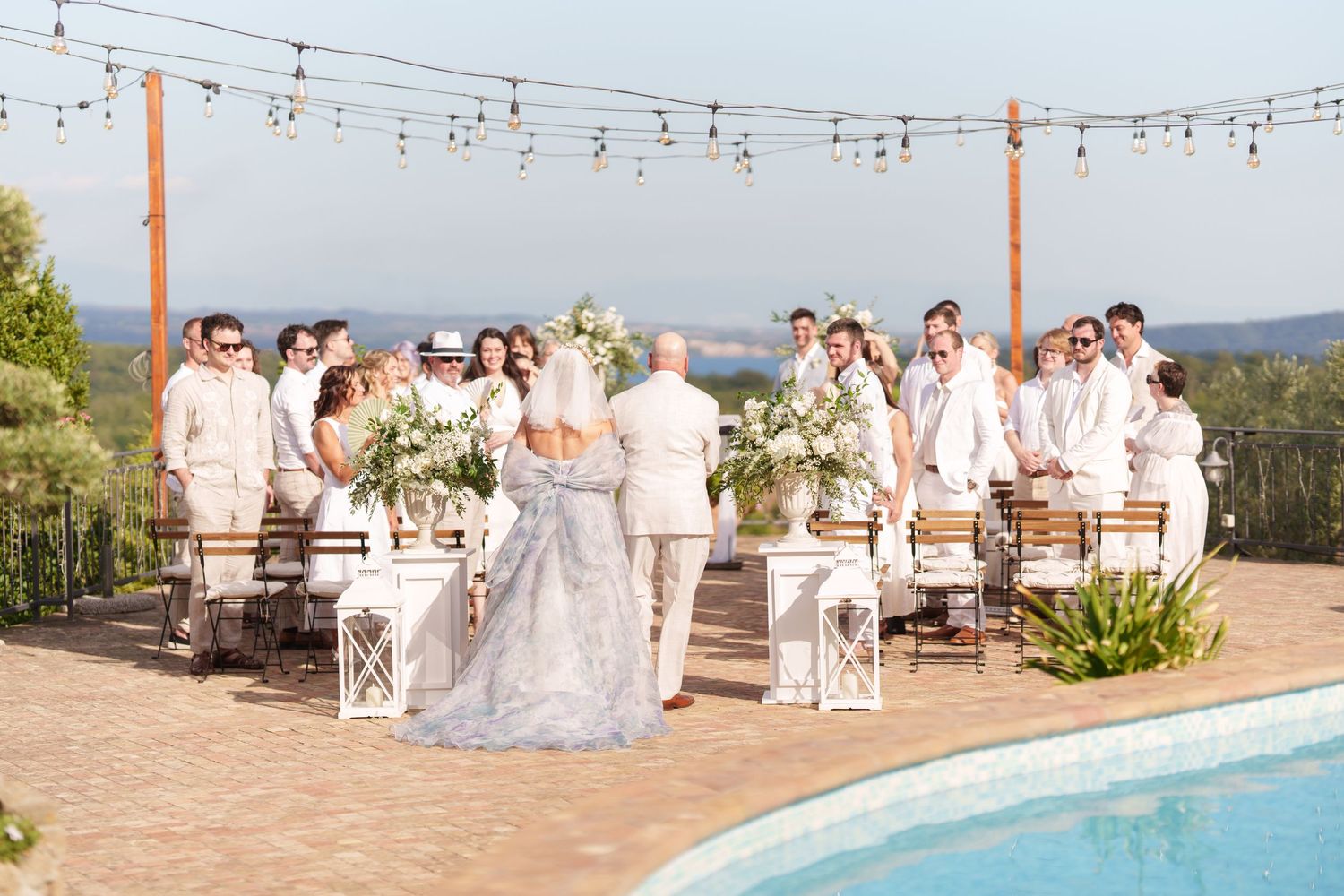 Outdoor wedding ceremony by a pool with guests in white attire, floral arrangements, and string lights under a clear sky.