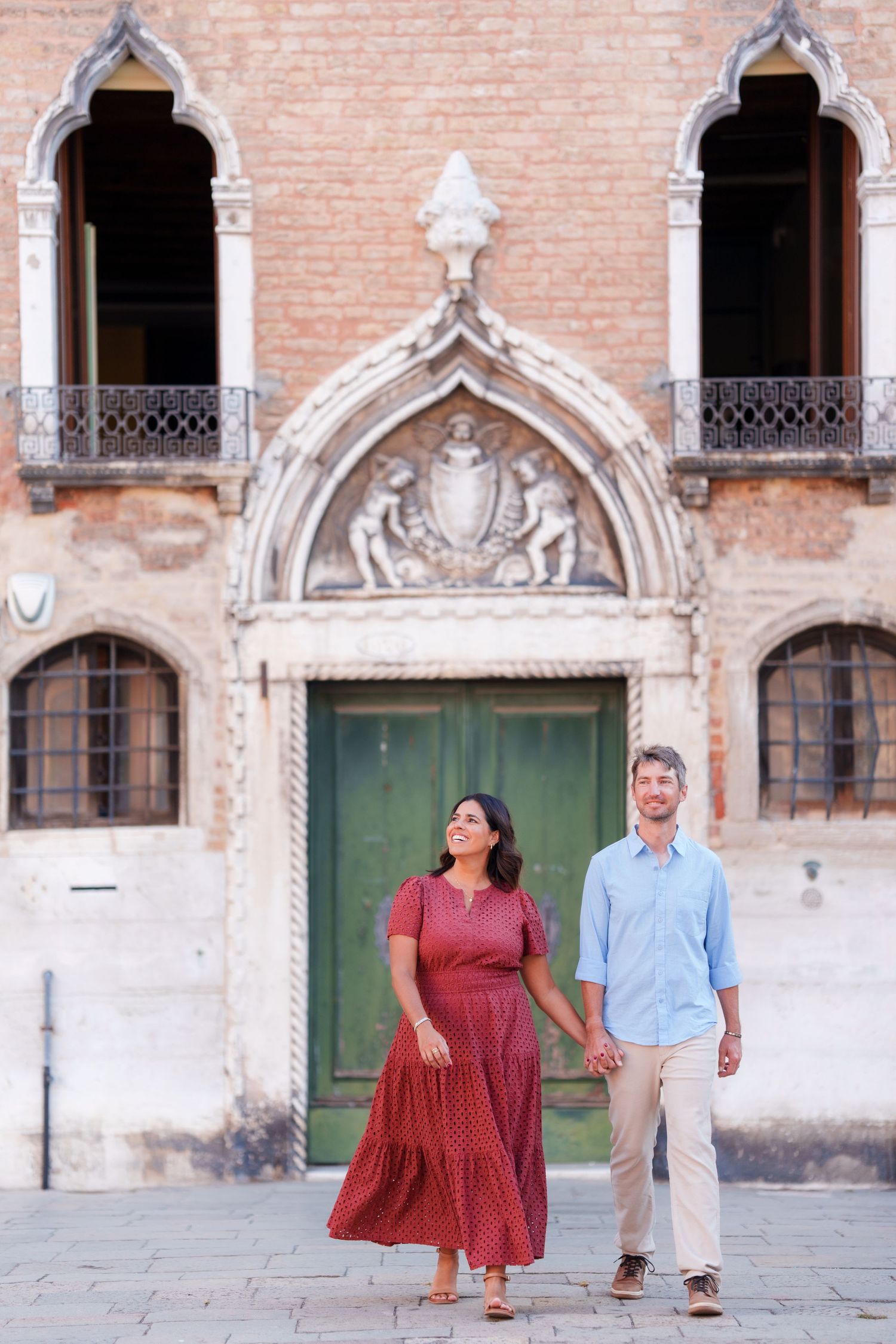 Couple holding hands walking in front of ornate Venetian Gothic building with green door in Venice, Italy.
