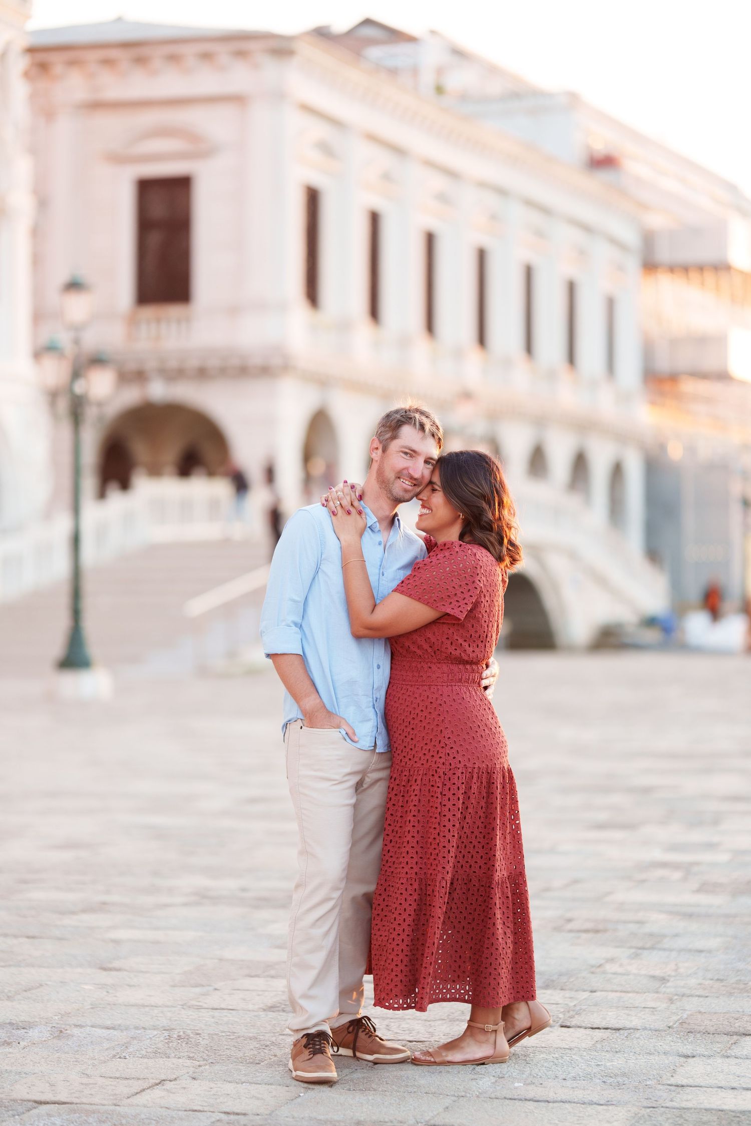 Couple embracing and smiling in a sunlit European plaza with historic architecture in the background.