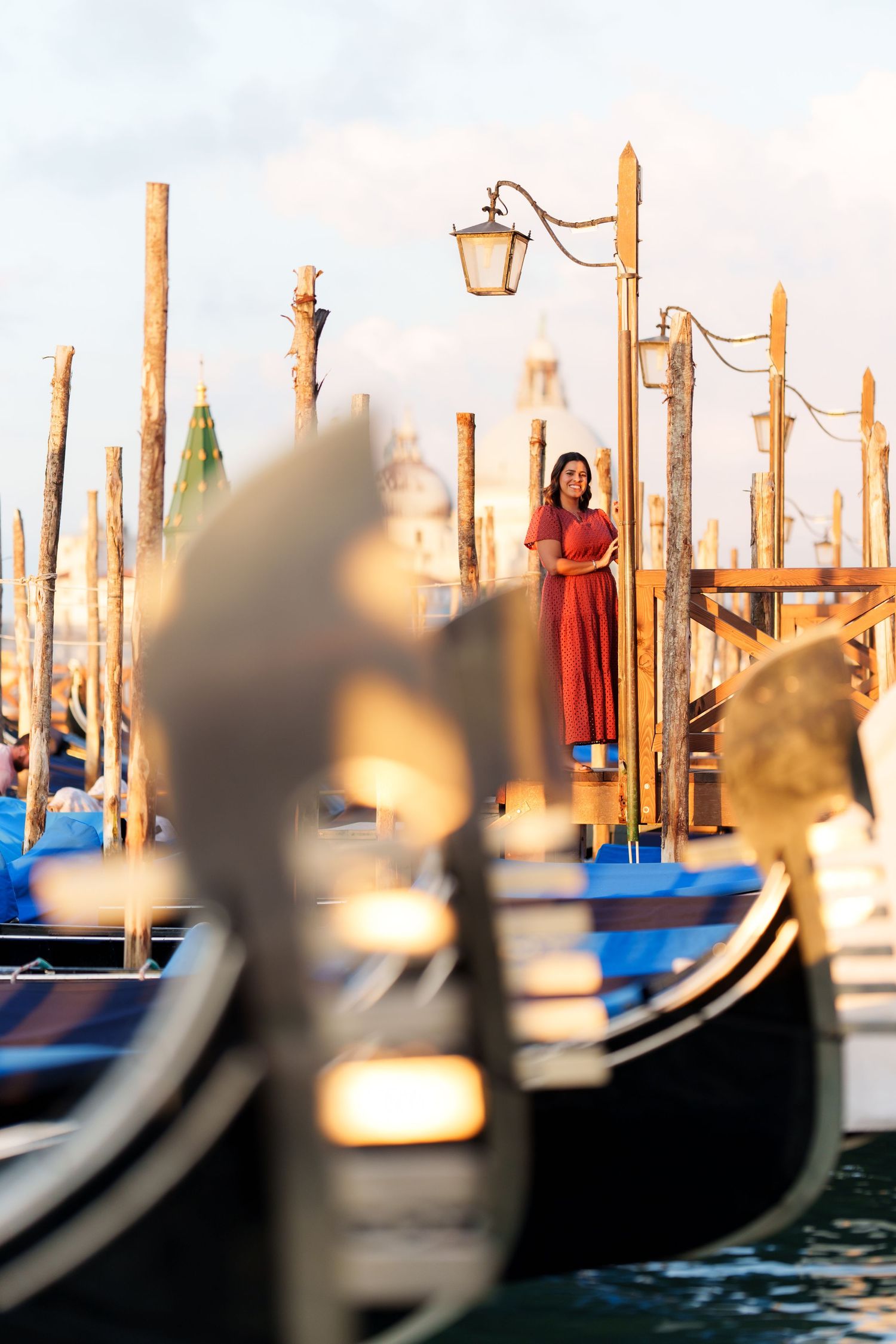 Woman in red dress standing on Venice gondola dock with wooden poles and street lamp at golden hour.