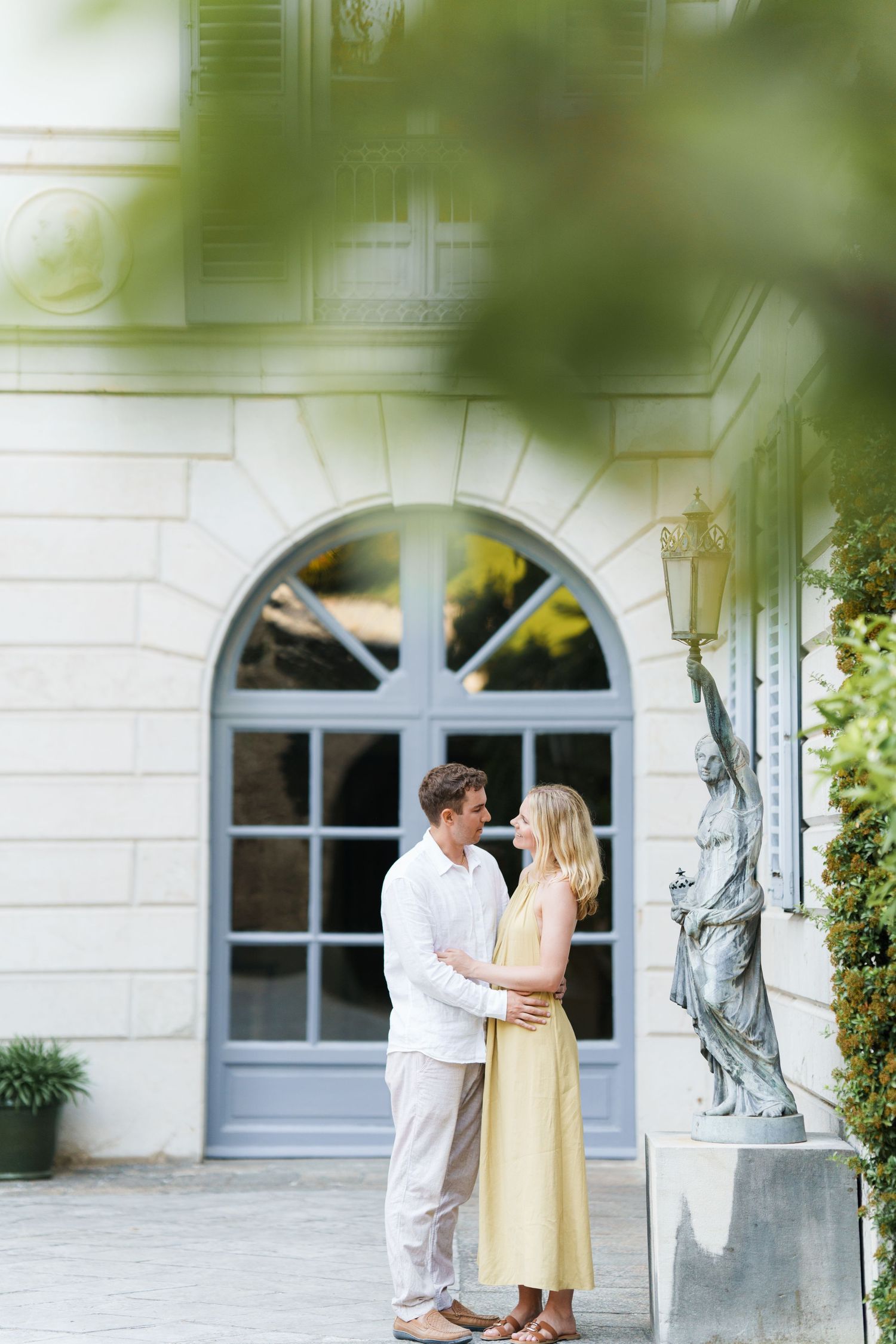 Couple embracing in front of elegant French architecture with arched windows and classical statue outdoors.
