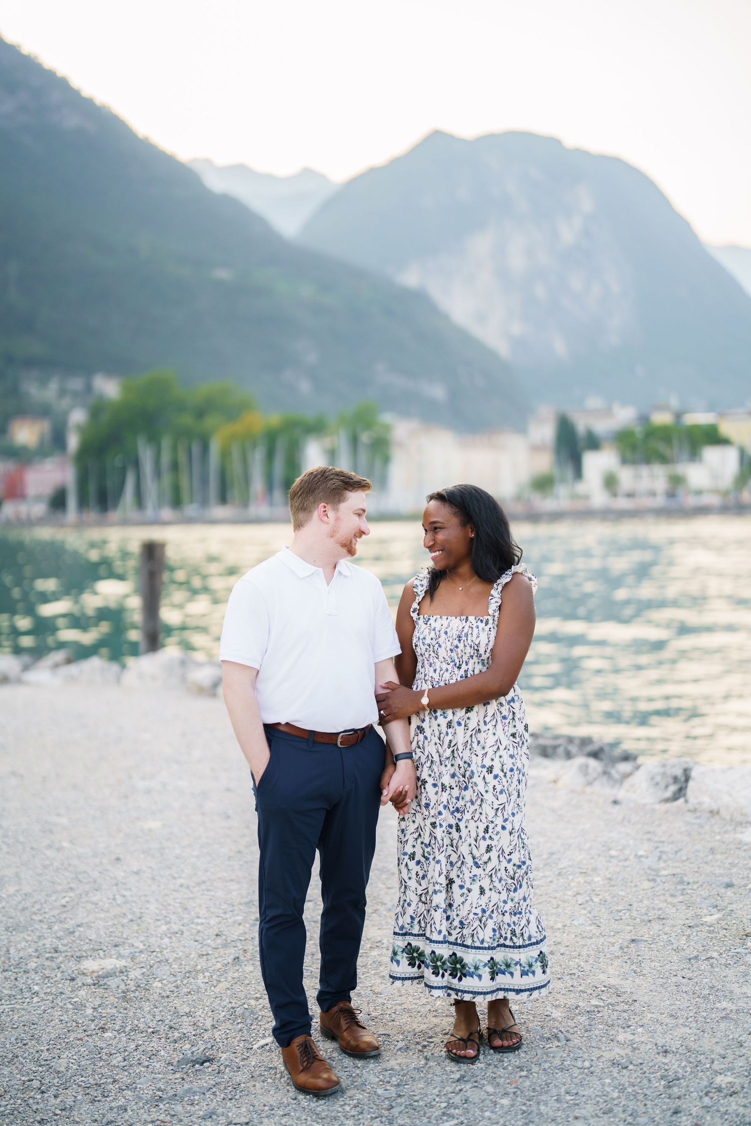 Happy interracial couple holding hands and smiling at each other by a scenic mountain lake.