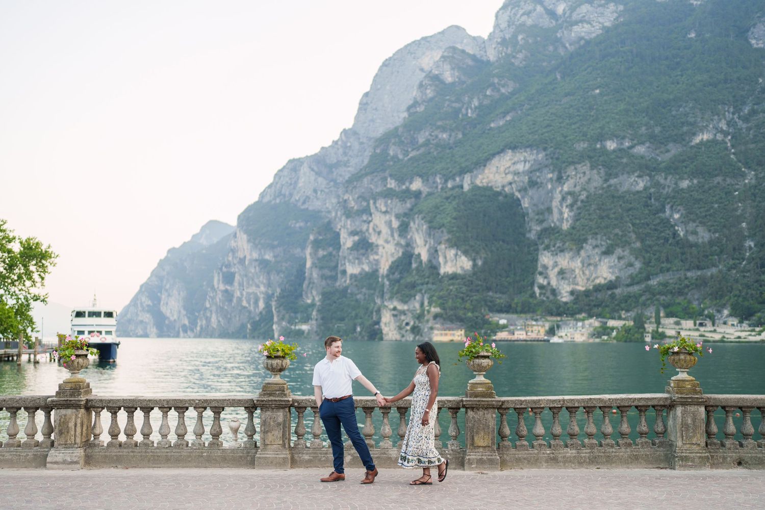 Couple walking hand in hand along a stone balustrade promenade with stunning alpine lake views behind them.