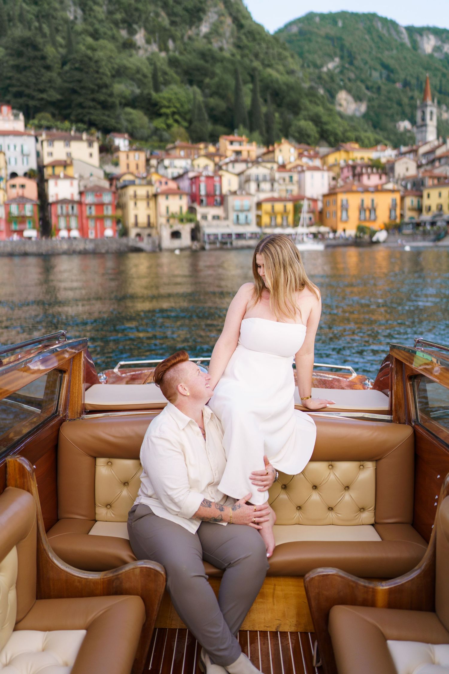 Couple on a luxury wooden boat at Lake Como, Italy, with colorful Varenna village buildings reflected in the water.