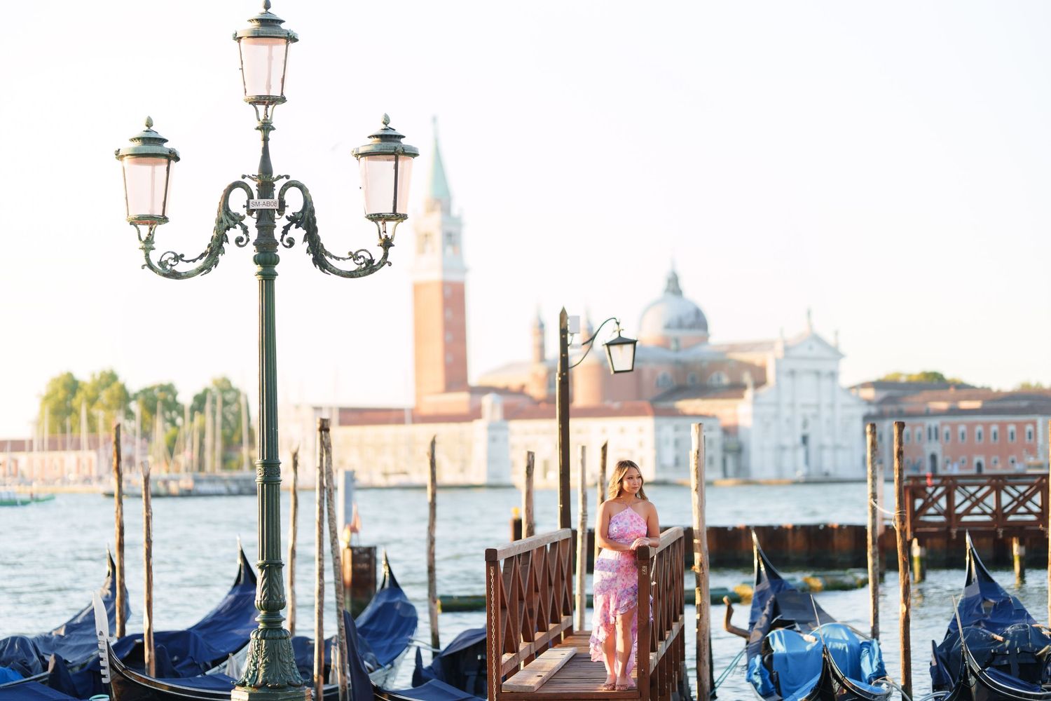 Woman in pink dress posing by Venice gondolas with San Giorgio Maggiore church in background.