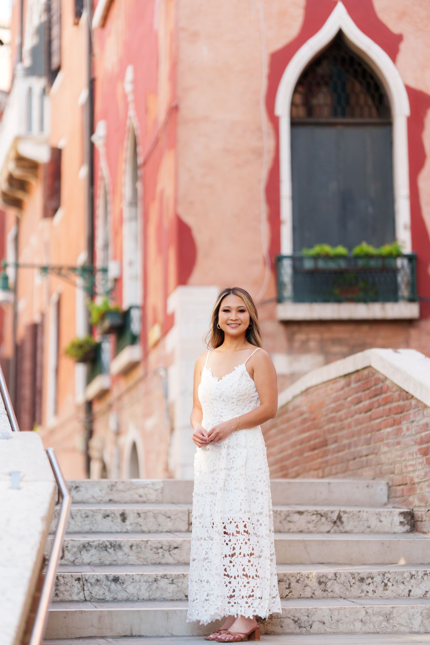 Woman in white lace dress smiling on stone steps in Venice, Italy, with colorful historic buildings behind her.