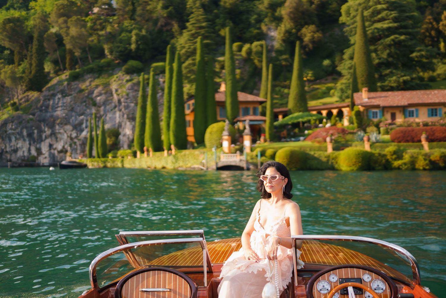 Woman in elegant dress seated on a wooden boat with Lake Como's lush gardens and cypress trees in background como lake bellagio