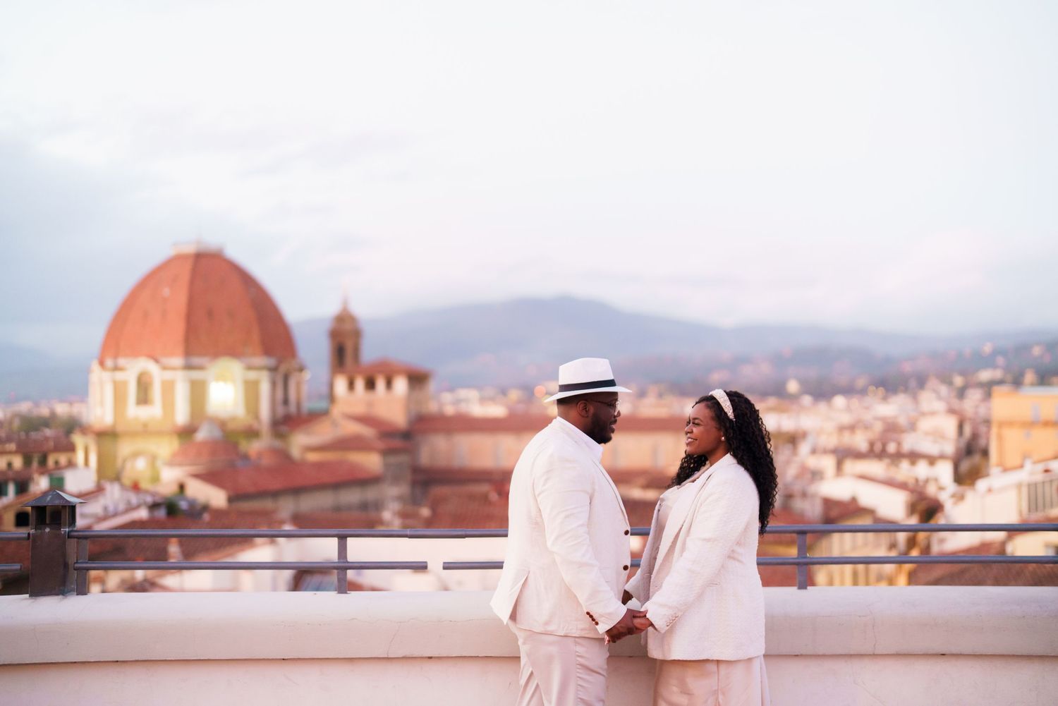 Couple in white attire holding hands on a rooftop in Florence, Italy, with the iconic Duomo dome in the background.