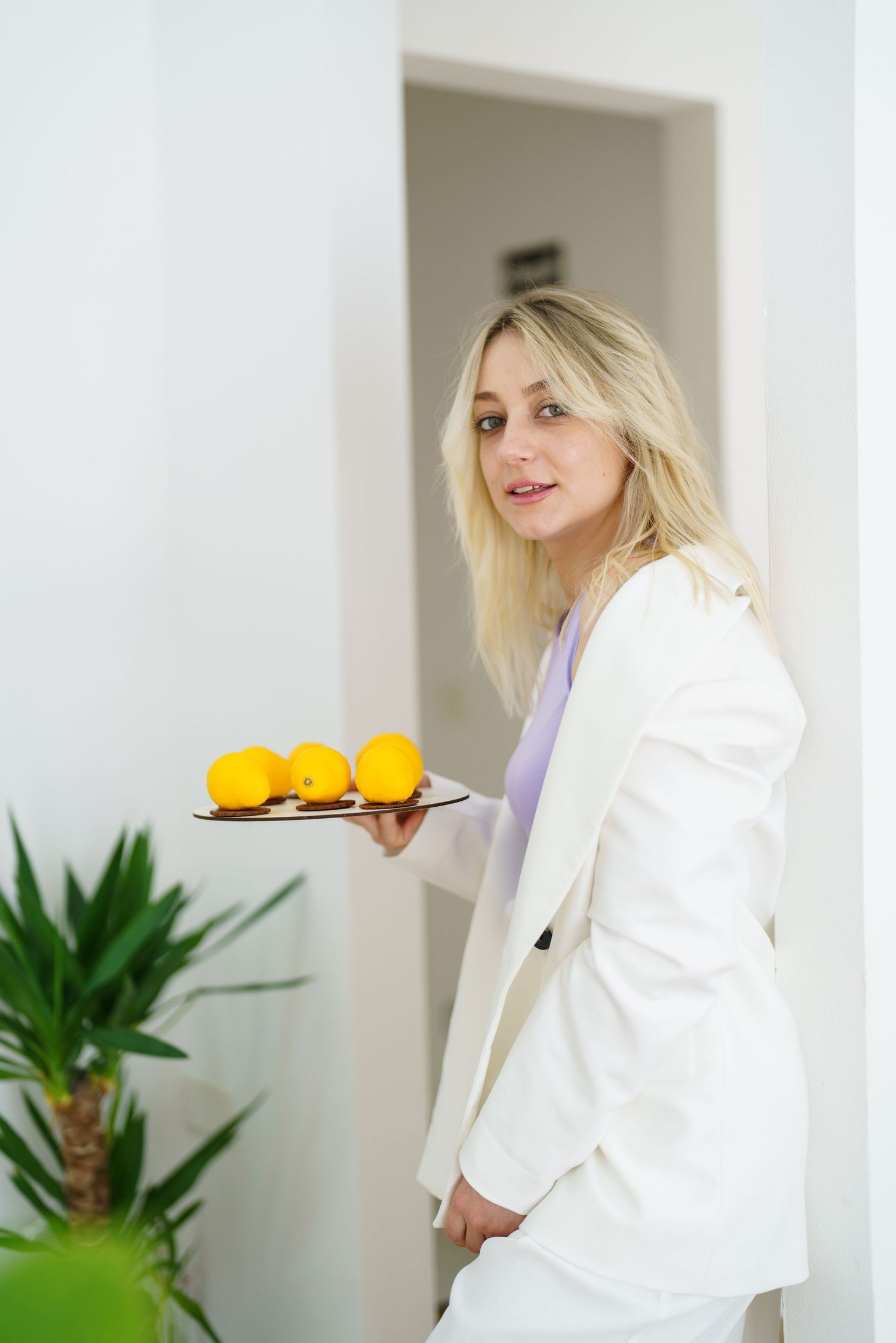 Blonde woman in white suit holding a tray of fresh oranges in a bright modern interior.