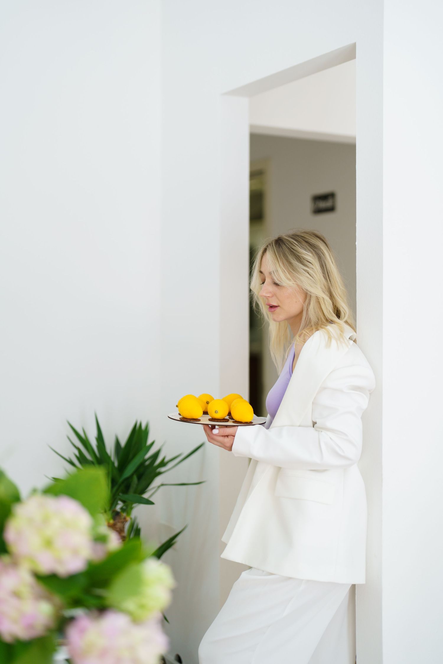 Blonde woman in white suit carrying a plate of yellow lemons through a bright white doorway with flowers in foreground.