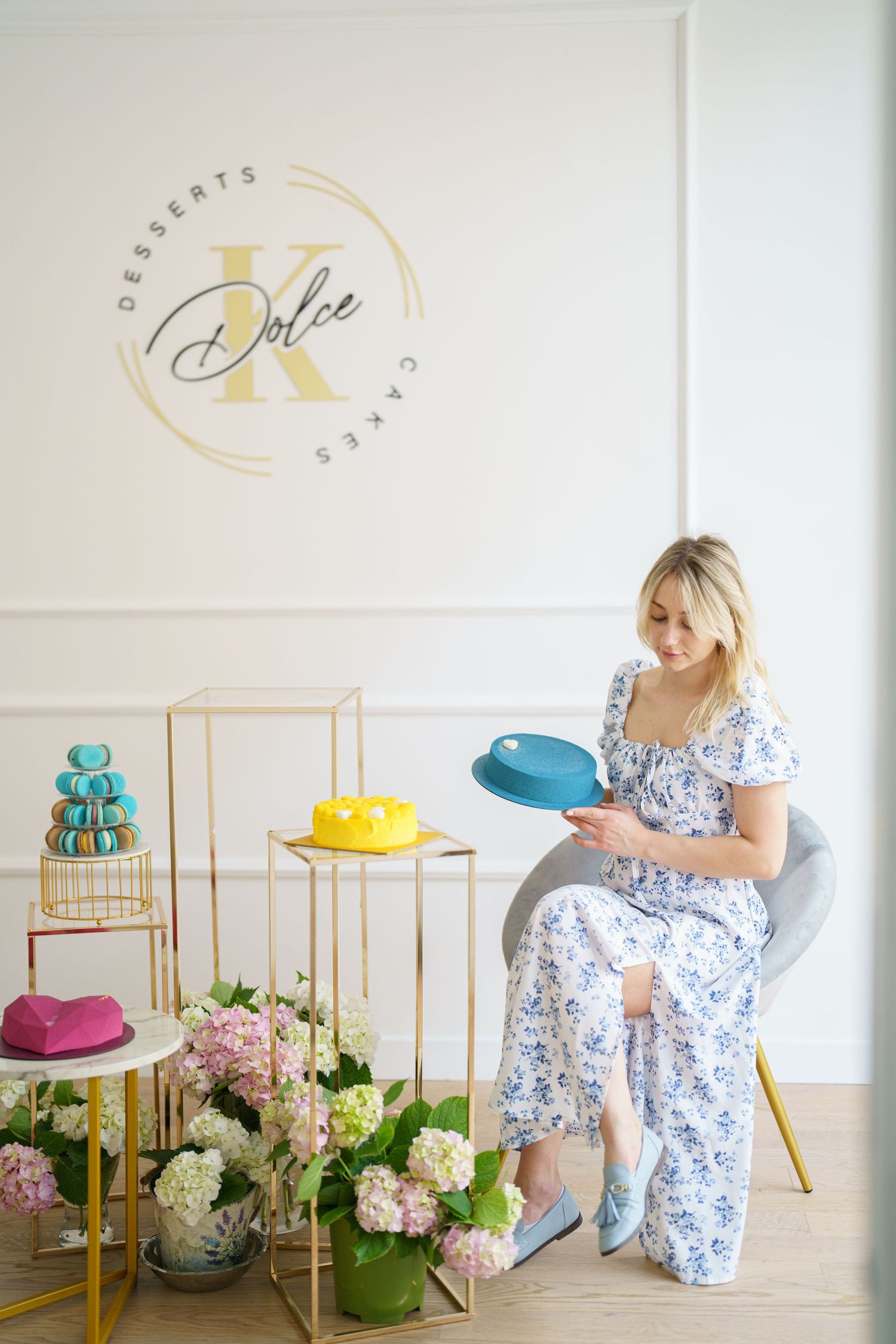 Woman in floral dress holding blue cake at Dolce K desserts shop with macarons and hydrangeas.