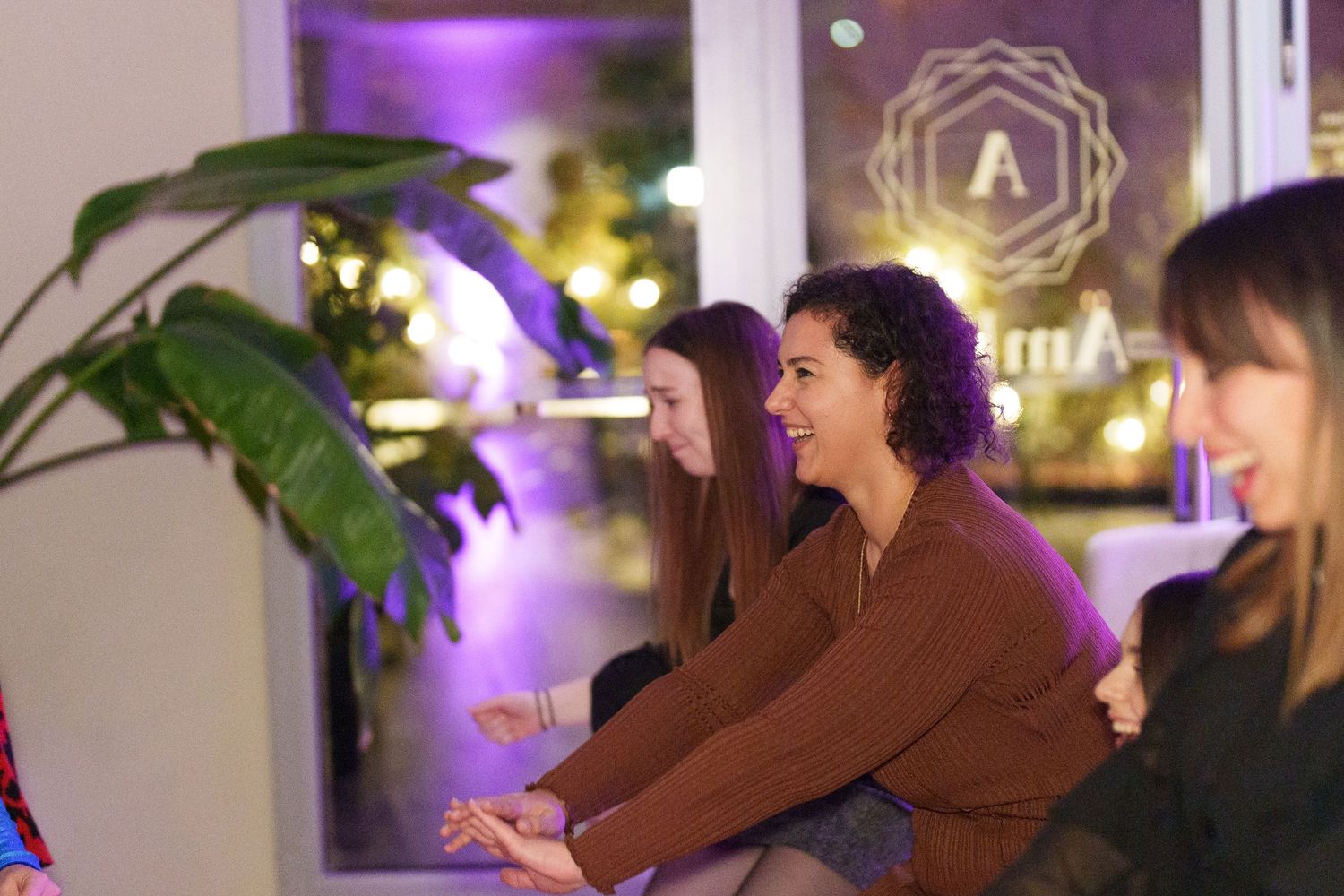Women smiling and socializing at a trendy purple-lit venue with plants and a geometric logo on the window.