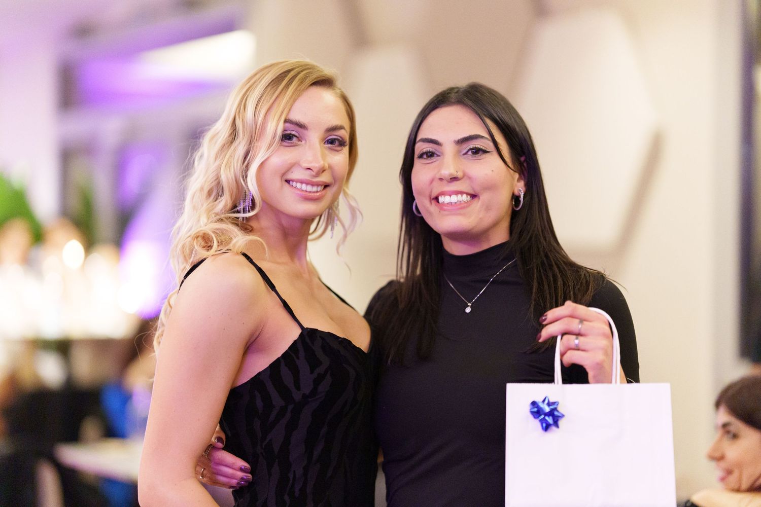 Two smiling women posing together at an event, one holding a white gift bag with blue ribbon.