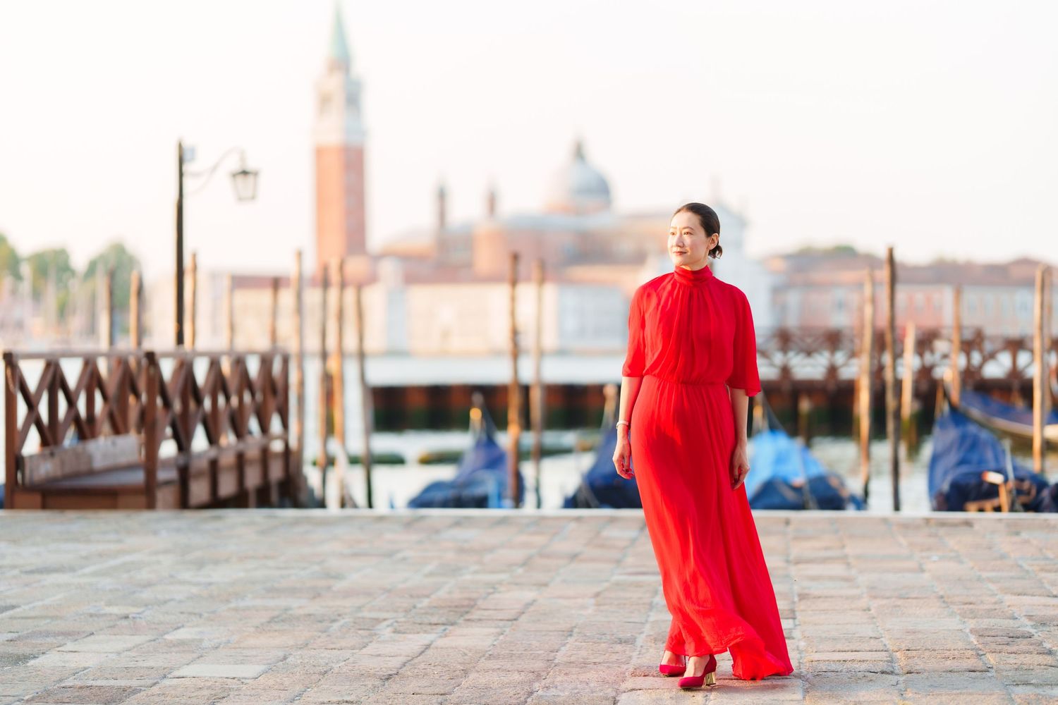 Woman in red maxi dress standing on Venice waterfront with gondolas and San Giorgio Maggiore in background.