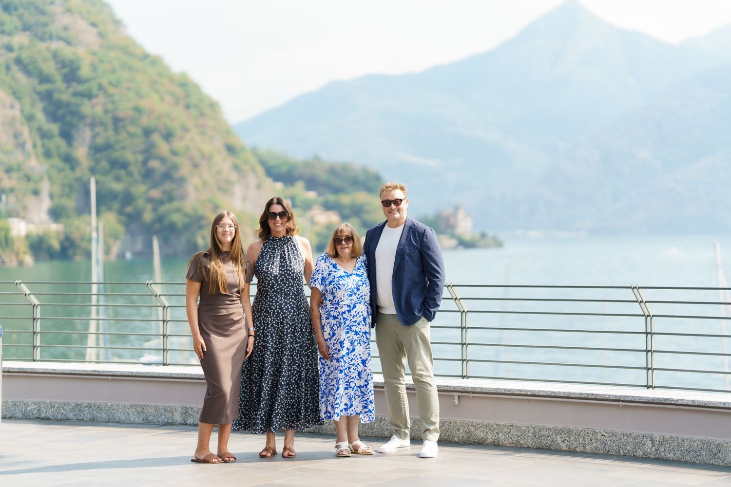 Four people posing together on a lakeside promenade with scenic mountain views in the background.