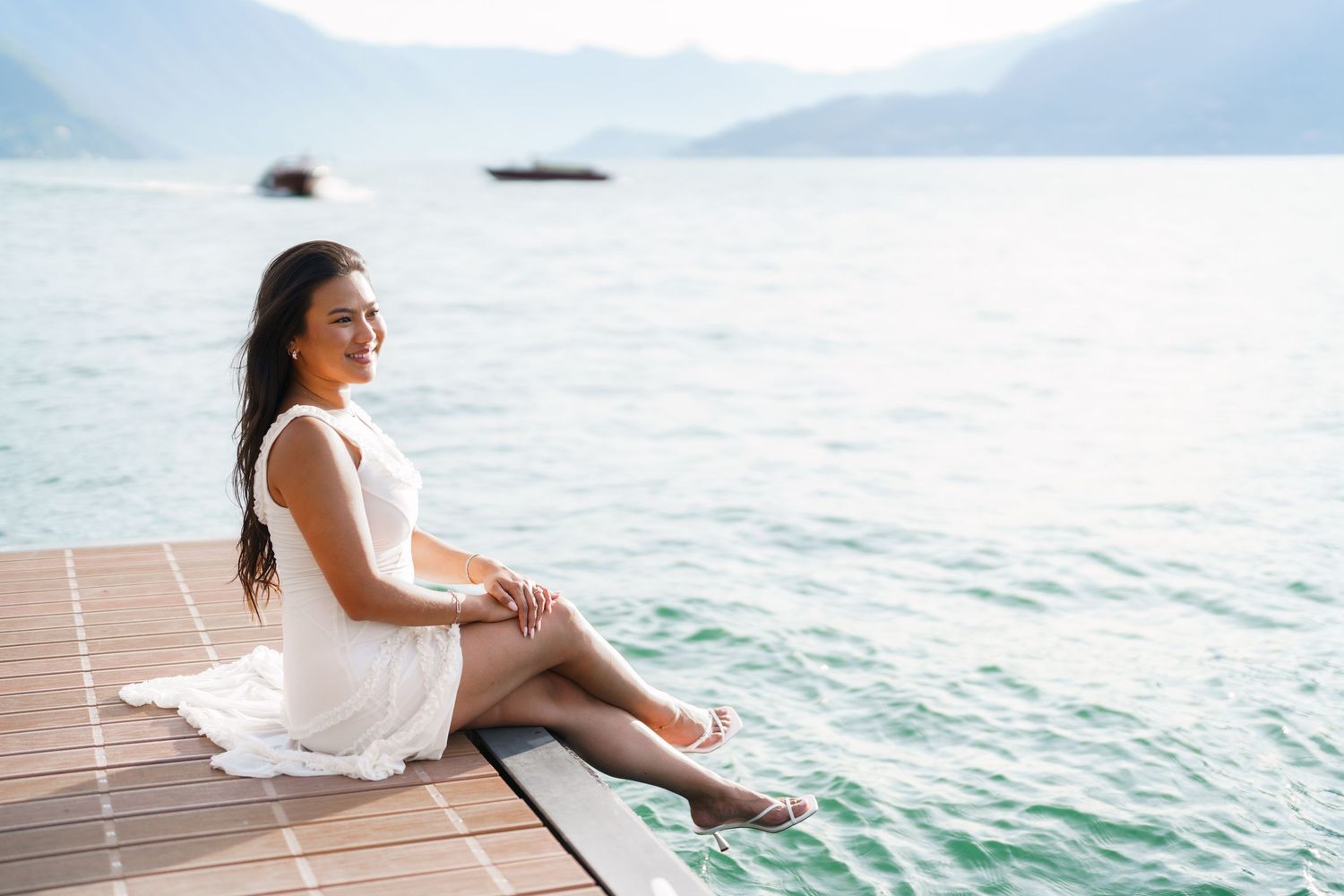 A smiling woman in a white dress sits on a wooden dock by a serene mountain lake on a sunny day.