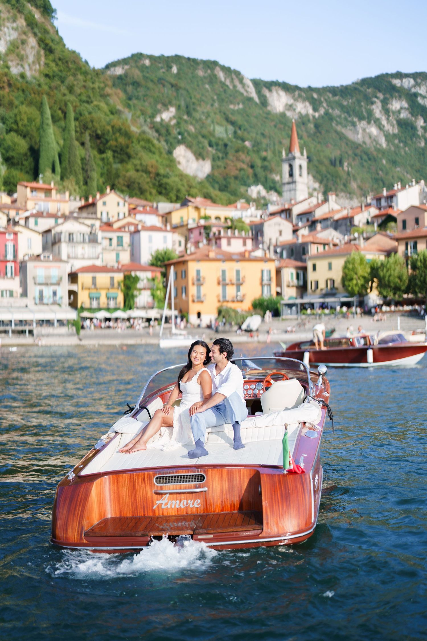 Couple embracing on a wooden motorboat named Amore on Lake Como with colorful Italian village in background.