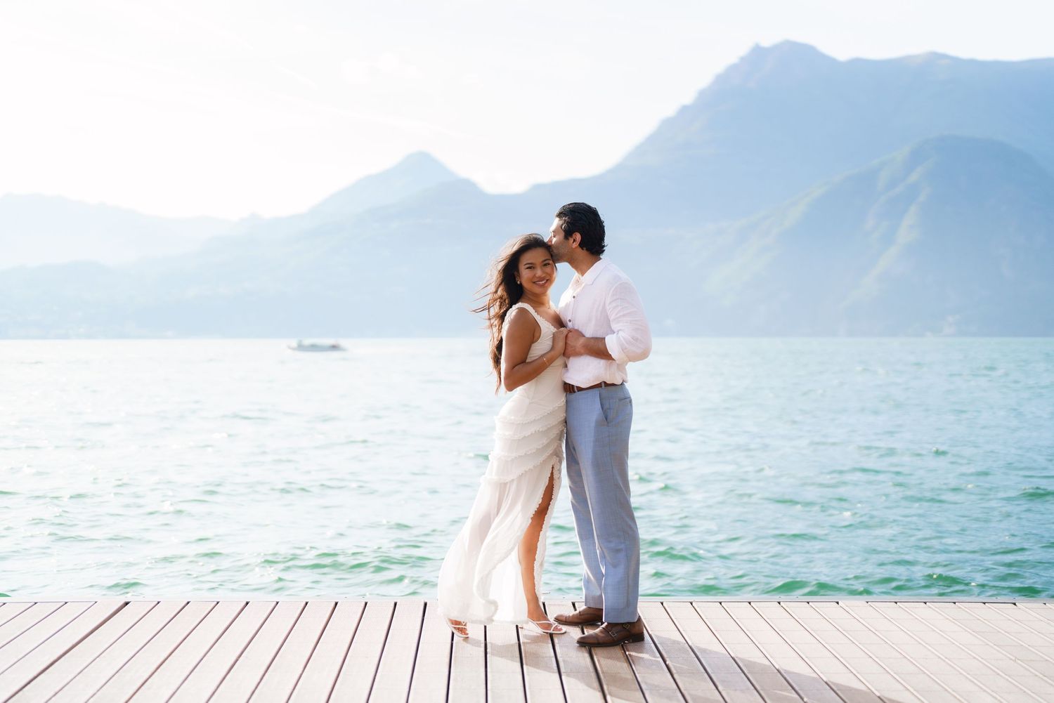 Couple embracing on a wooden dock with turquoise lake and mountain backdrop on a sunny day.