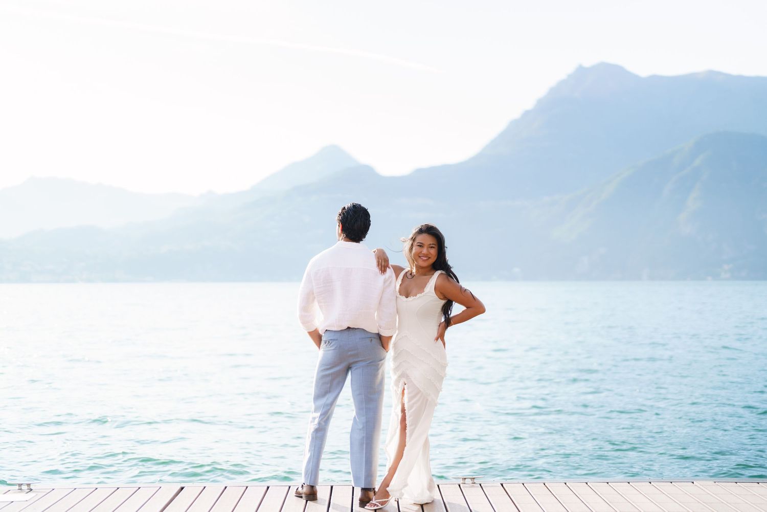 Couple in white outfits standing on a dock overlooking a serene mountain lake on a sunny day.