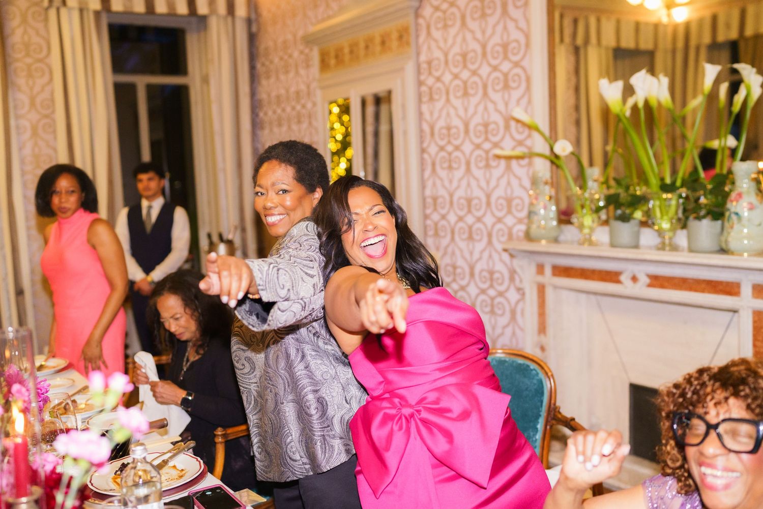 Two women in elegant attire laugh and dance together at a festive indoor celebration with guests in background.