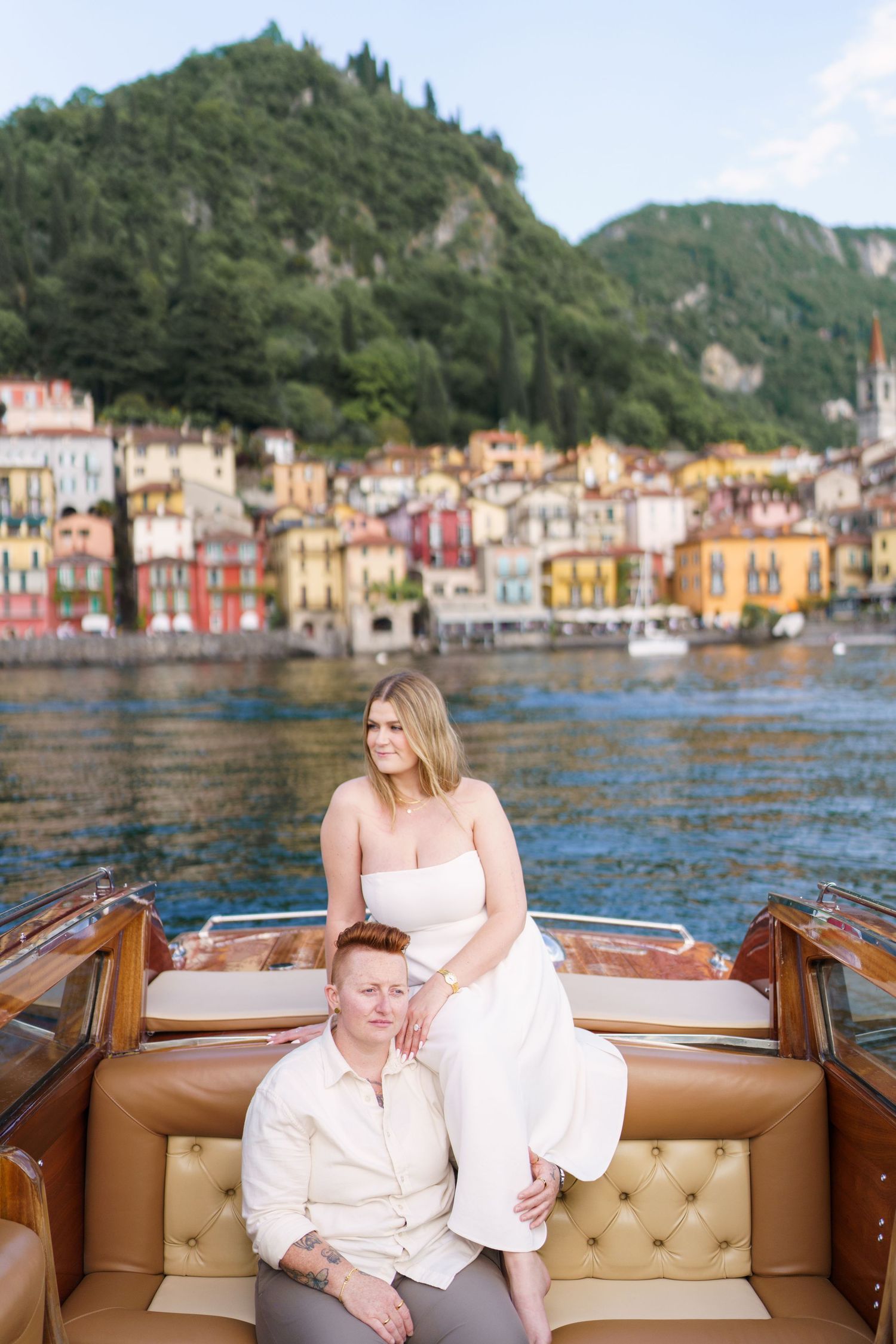Couple posing on a luxury wooden boat with colorful Italian lakeside village and green hills in the background.