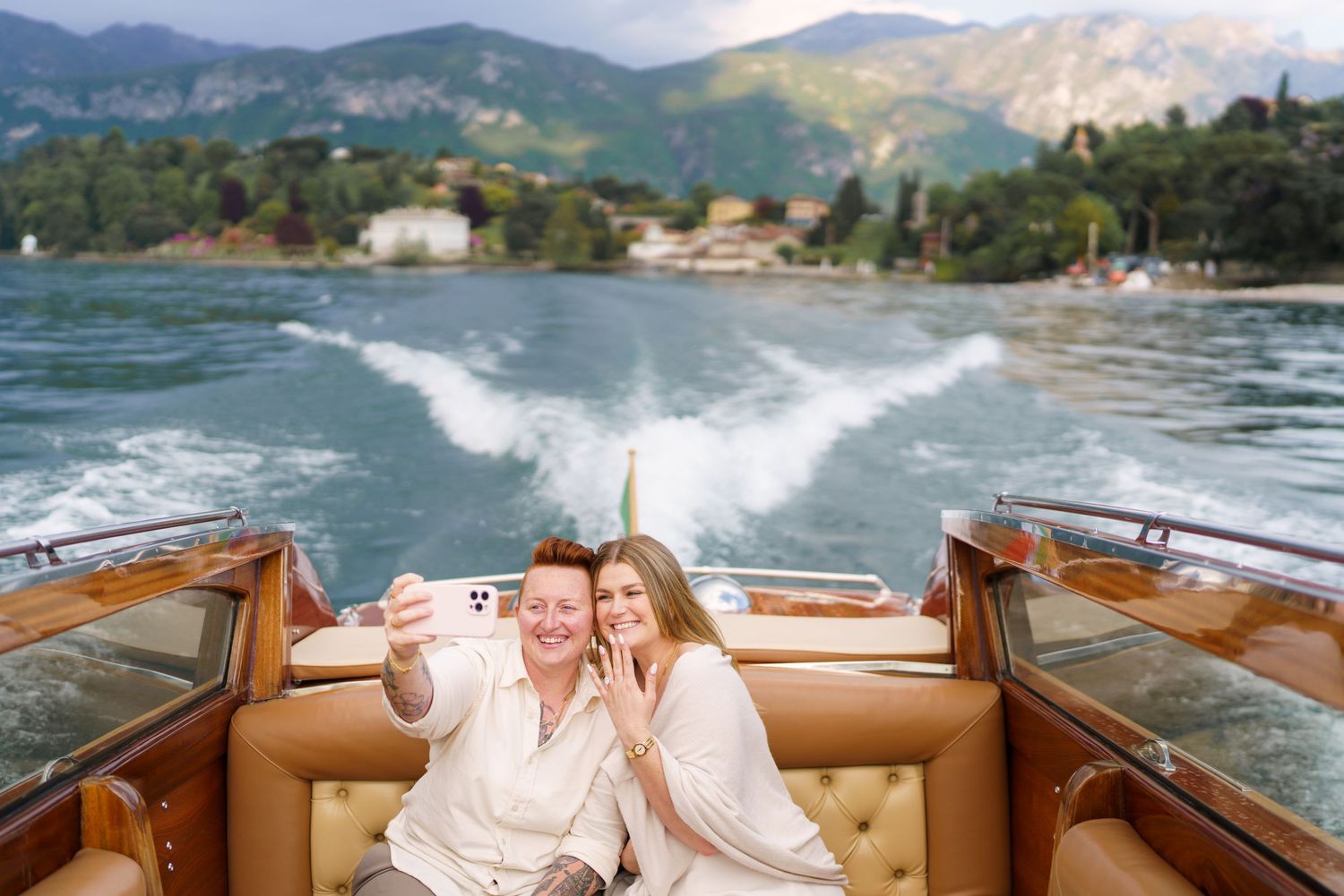 Two women take a selfie on a luxury wooden speedboat on a scenic Italian lake with mountains behind.