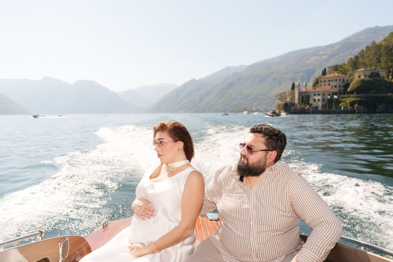 Pregnant couple enjoying a scenic boat ride on Lake Como, Italy, with mountains and a villa in the background.