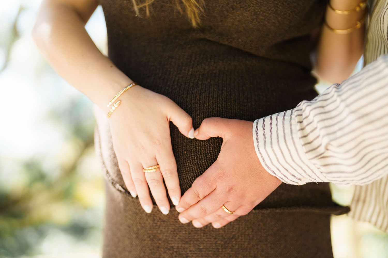 Couple forming a heart shape with their hands on a pregnant belly, wearing gold rings and bracelets.