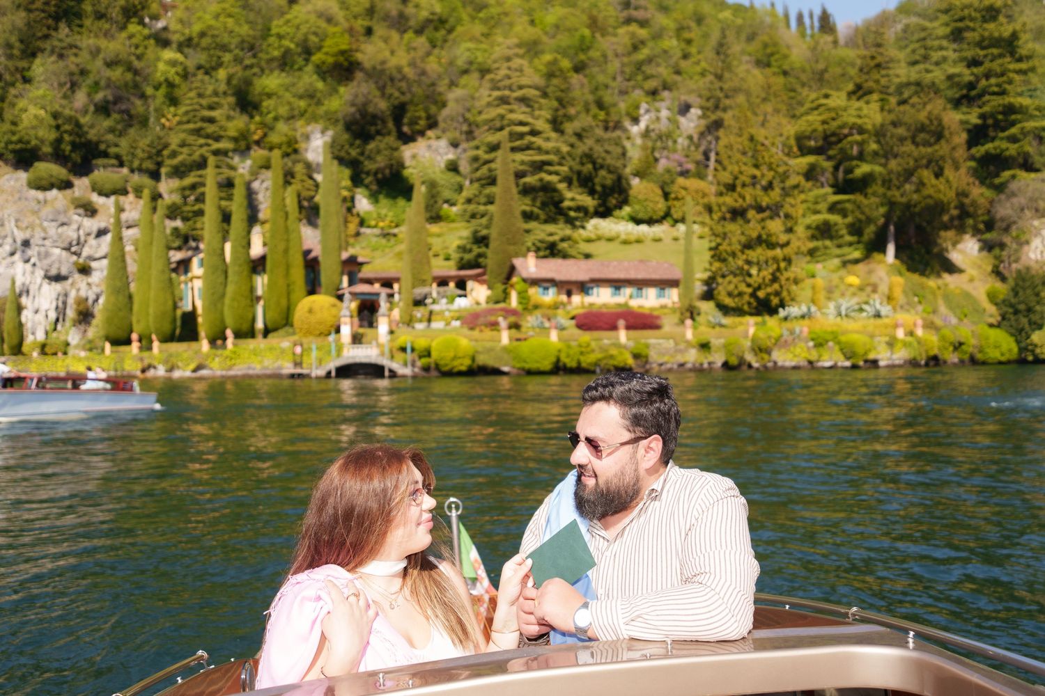 Couple enjoying a boat ride on a scenic lake with lush green hills and a charming villa in the background.
