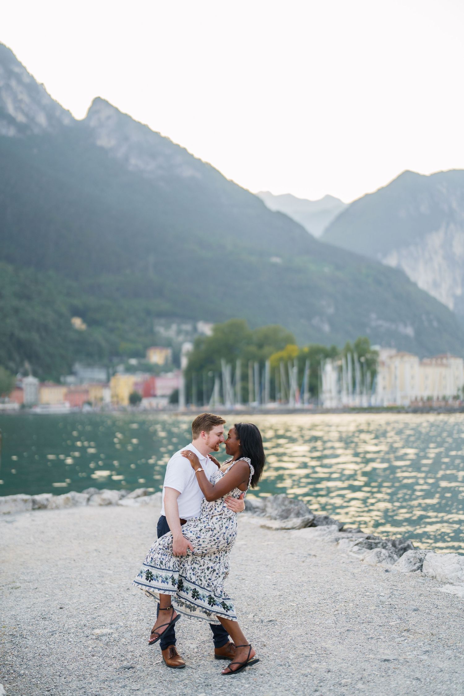 Interracial couple sharing a romantic kiss by a lakeside with mountains in the background.