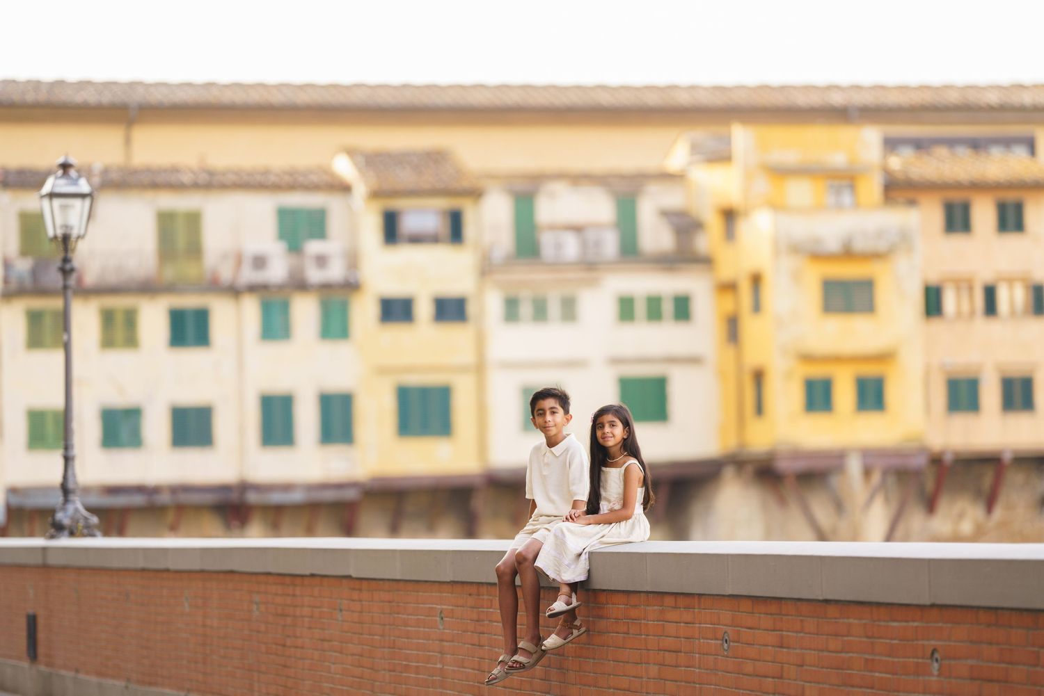 Two children in white outfits sit on a wall with colorful Ponte Vecchio buildings in Florence, Italy behind them.