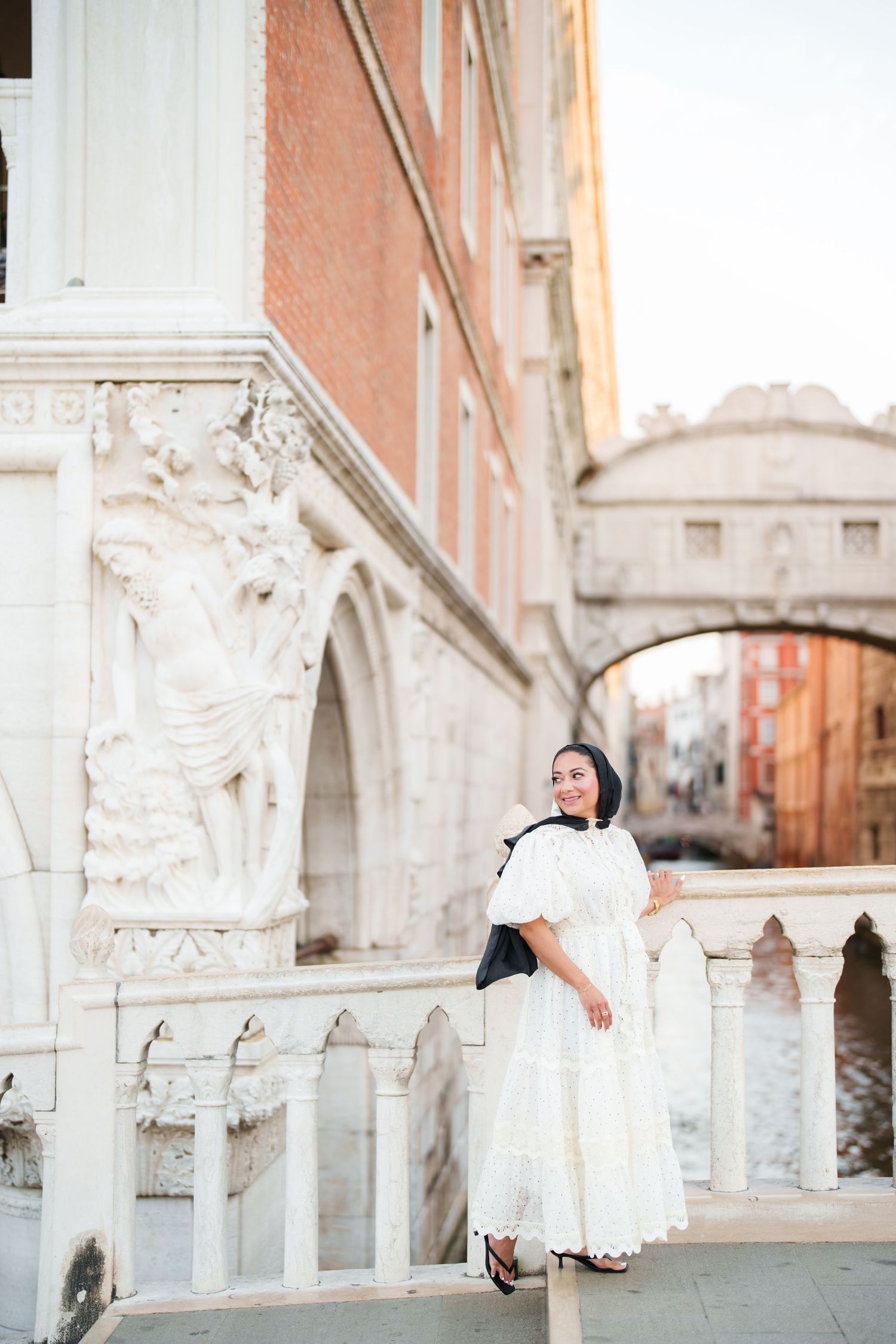 Woman in white dress posing near ornate marble columns with Venice's Bridge of Sighs in the background.