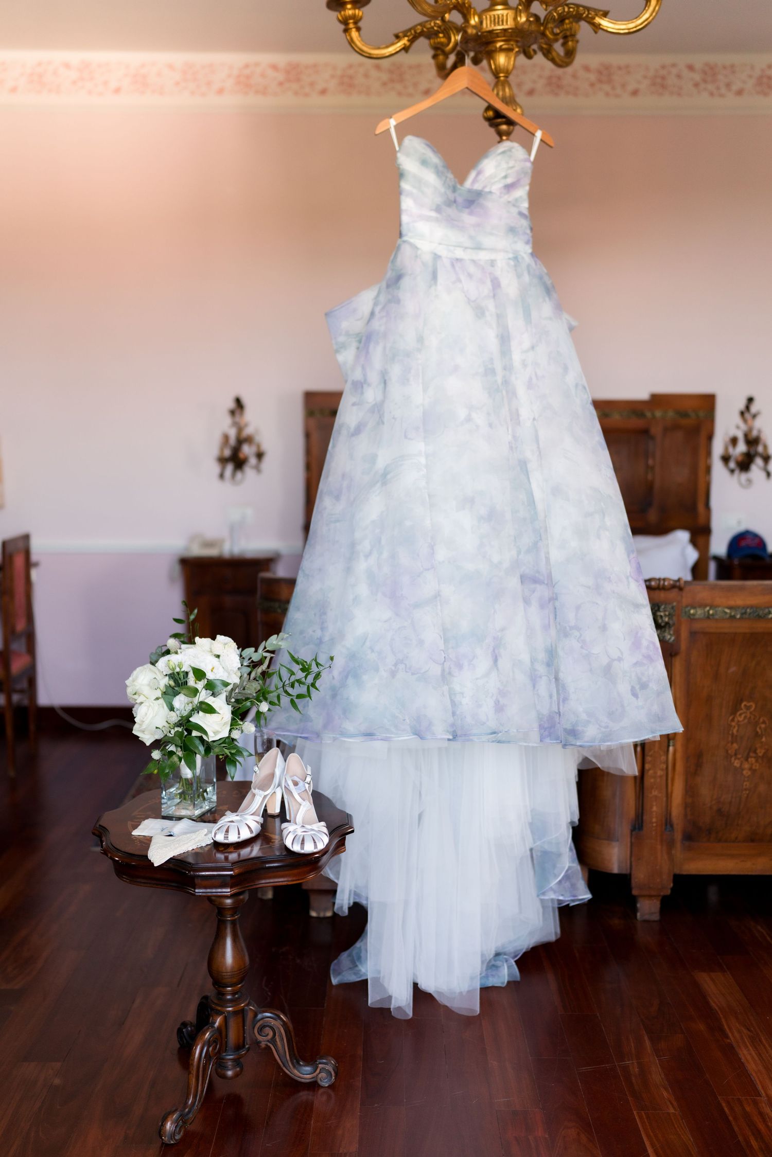 A floral blue and white wedding gown hanging from a chandelier beside bridal shoes and bouquet in an elegant room.