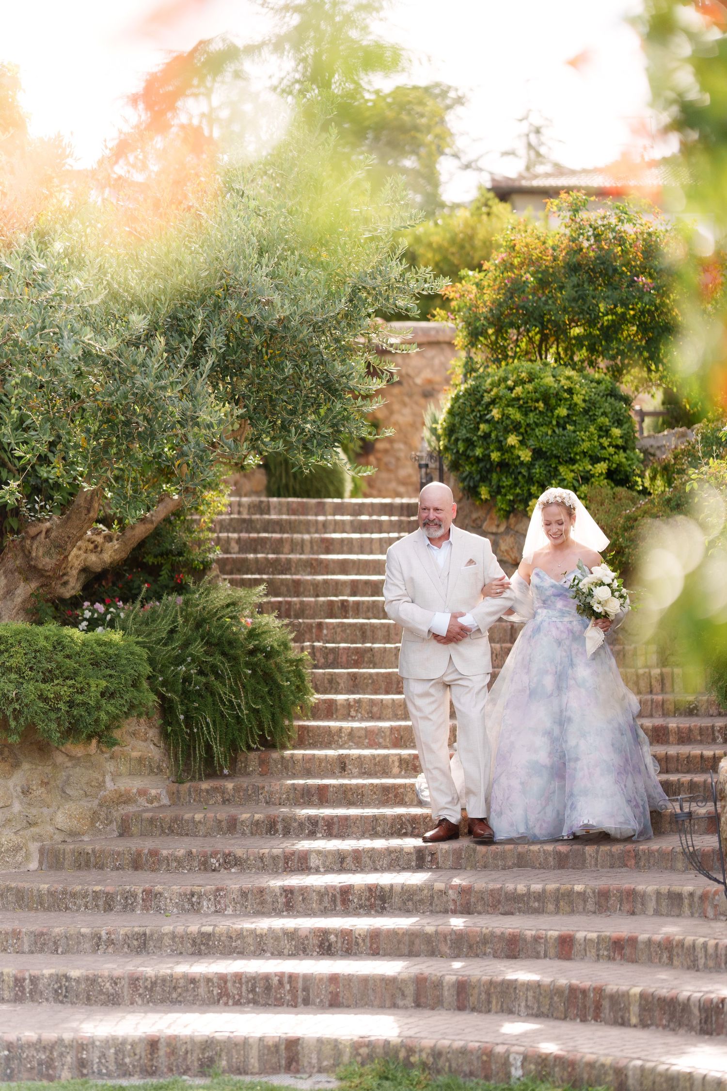 Bride in lavender gown walks down stone garden steps with her father on a sunny wedding day.