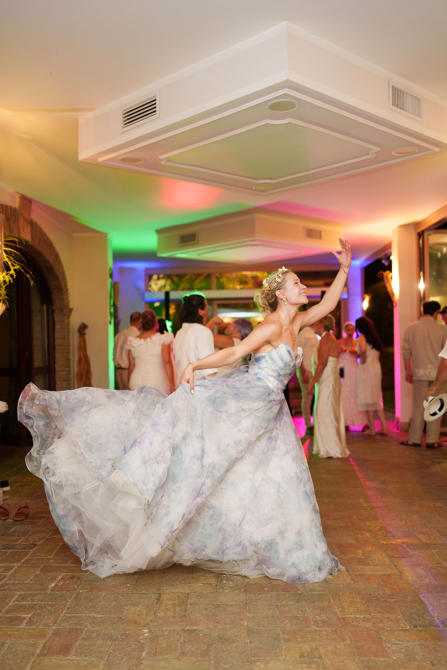 Bride in floral gown twirling on dance floor at wedding reception with colorful lights.