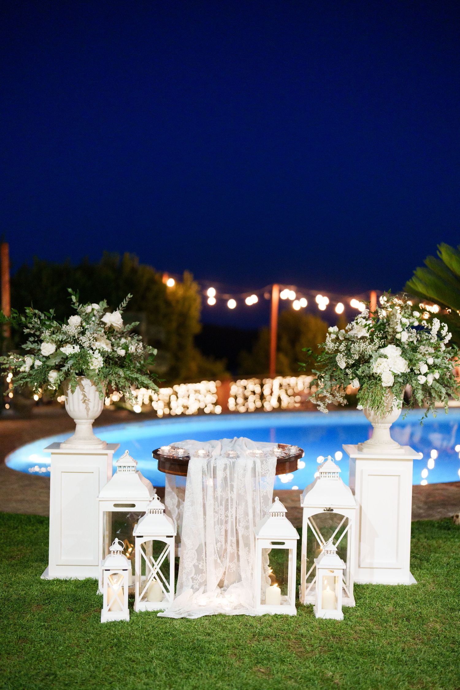 Elegant outdoor wedding setup at night with white lanterns, floral arrangements, and a glowing pool in the background.