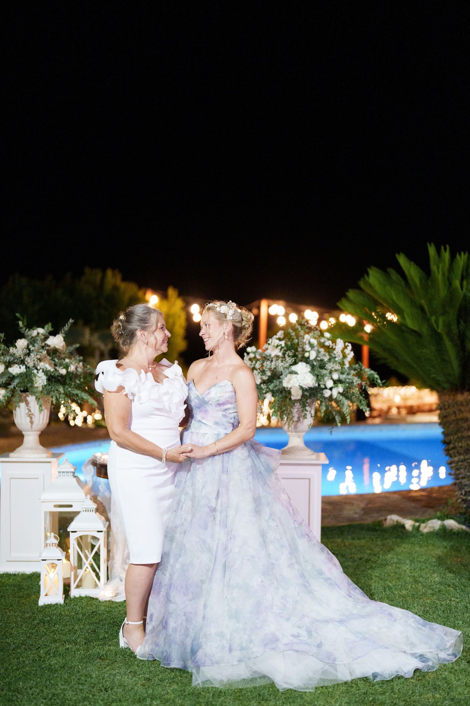 Two brides in white and lavender gowns hold hands by a lit pool at a night wedding ceremony.