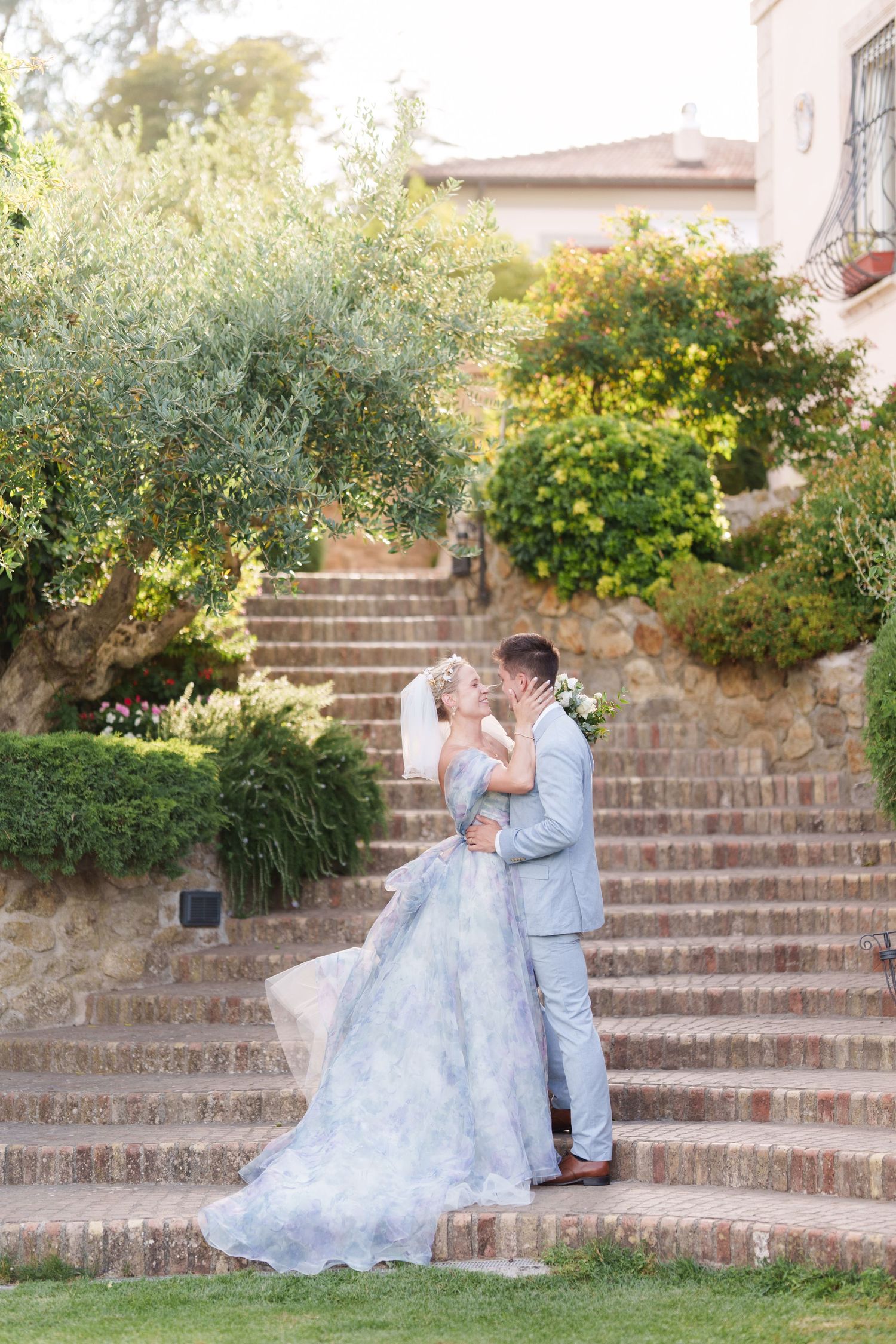 Bride in blue gown and groom in gray suit share a kiss on stone steps surrounded by lush greenery.