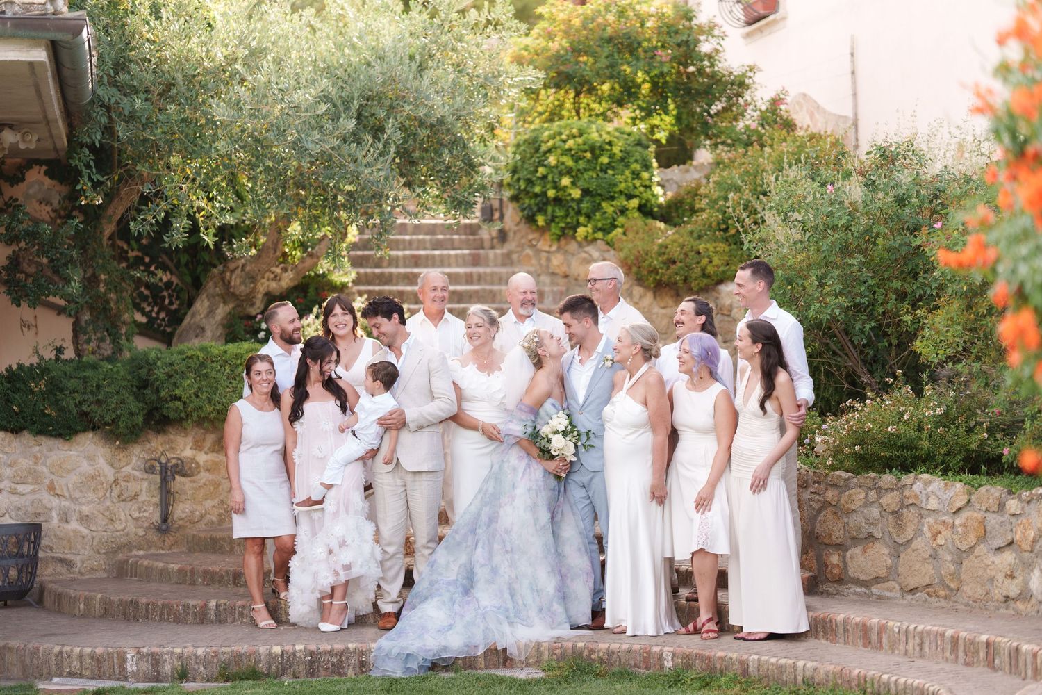 Wedding party in white attire poses on stone steps surrounded by lush greenery at an outdoor ceremony.