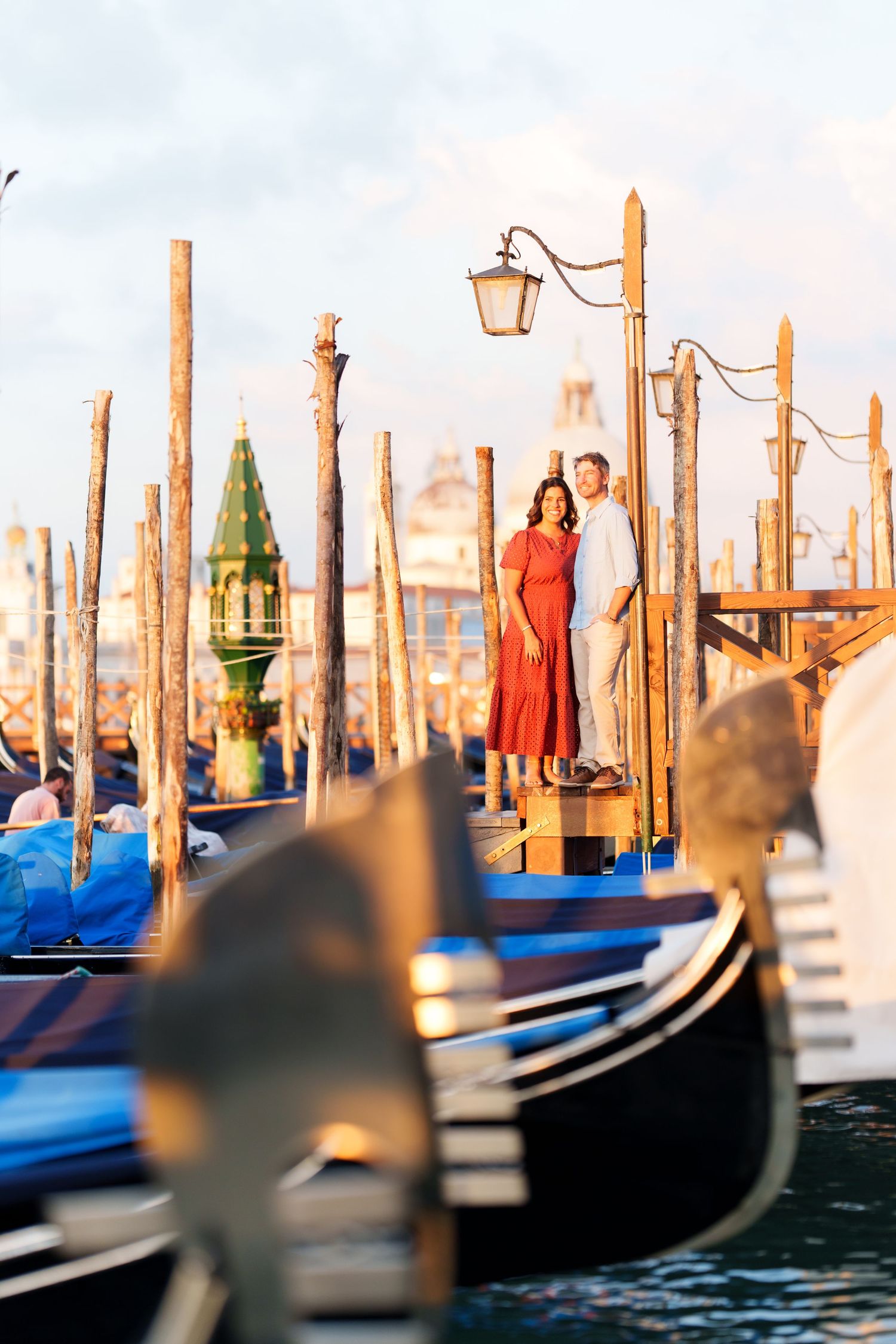 Romantic couple standing on a Venice gondola dock at golden hour, with iconic gondolas and wooden poles in the foreground.