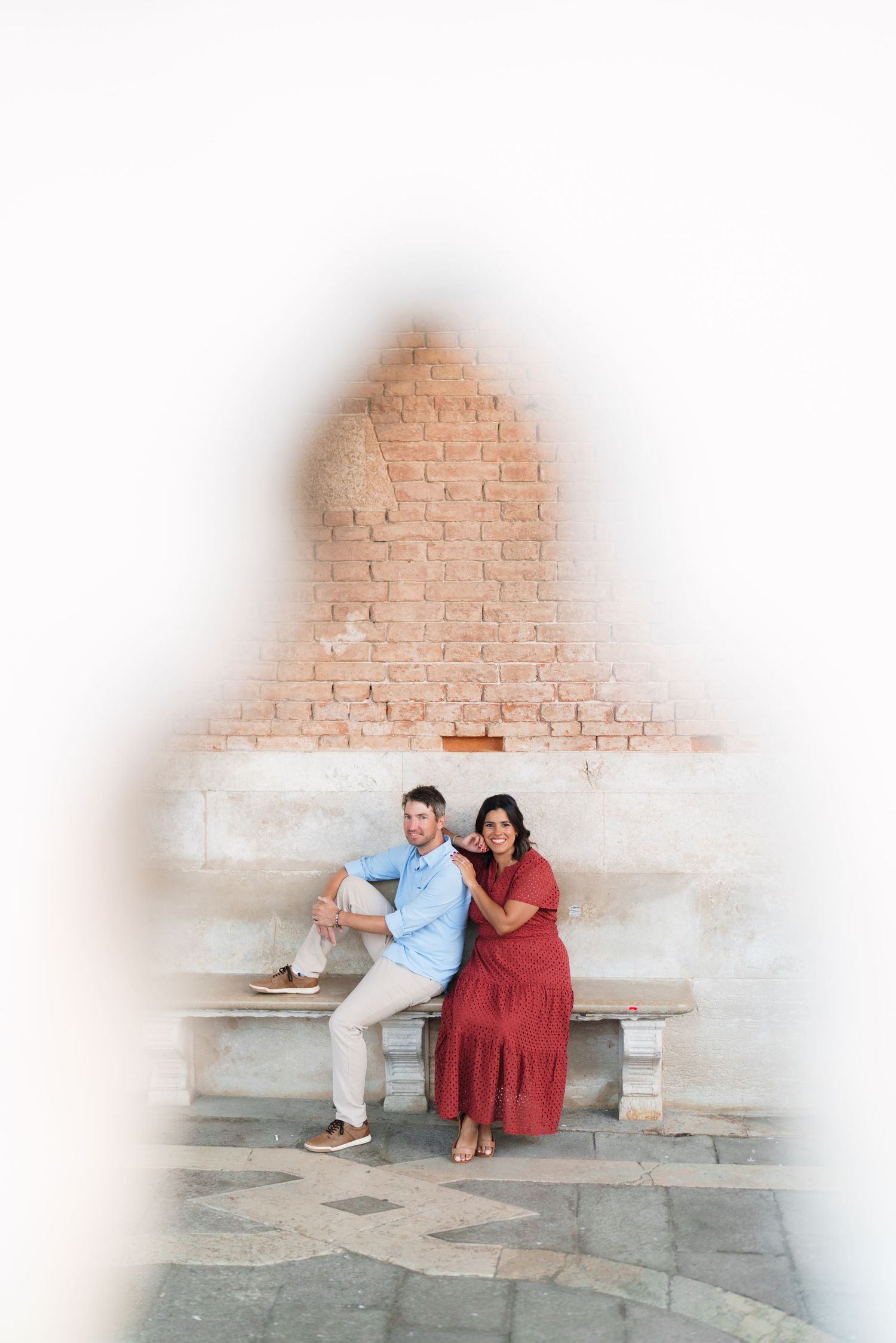Couple sitting on a stone bench in Venice, framed by a keyhole arch, with a brick wall backdrop.
