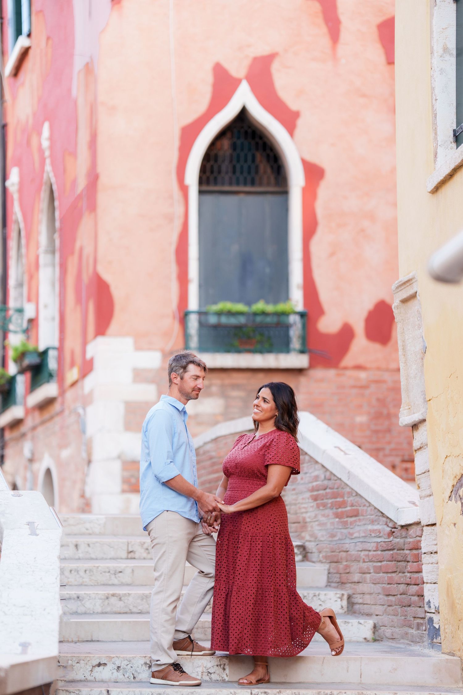 A couple holds hands and smiles on Venice steps, with colorful Italian architecture in the background.