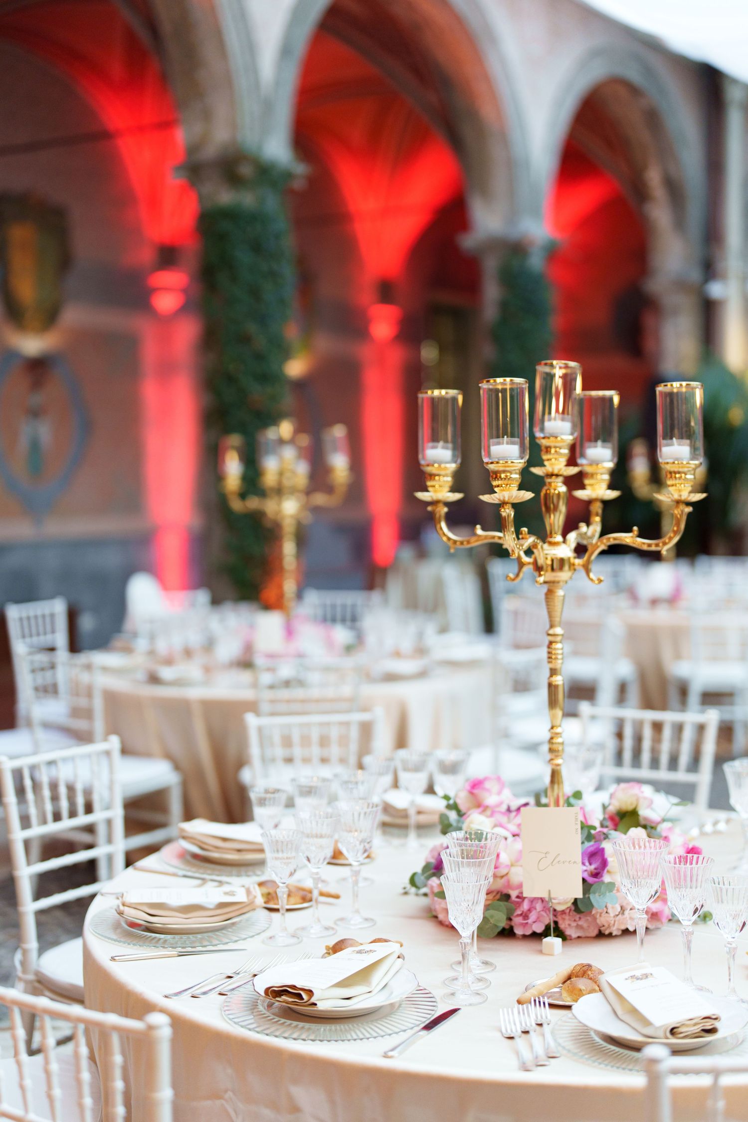 Elegant wedding reception tables with gold candelabras and red uplighting in an ornate venue.